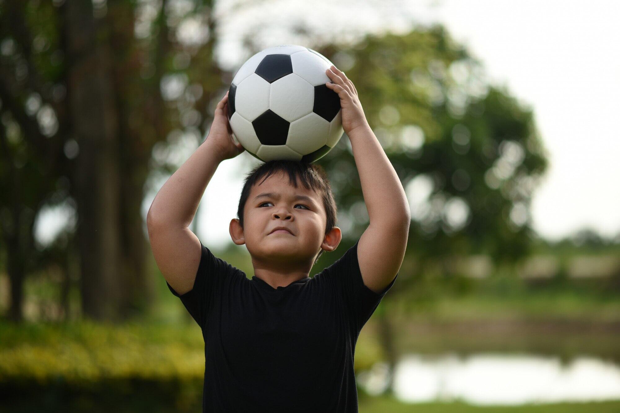 niño pequeño sosteniendo una pelota de futbol con las dos manos sobre su cabeza