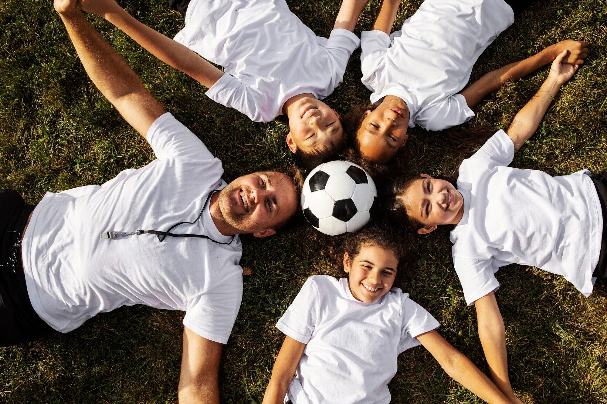 4 niños junto a su entrenado echados en la cancha sonrientes con una pelota de futbol en medio de la foto