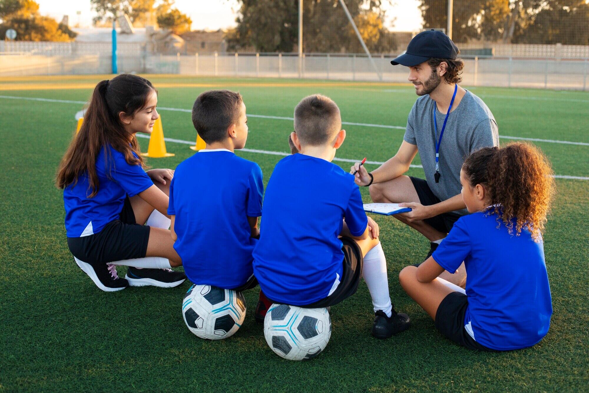 4 niños sentados en la cancha con sus balones de futbol junto a su entrenador explicando la clase