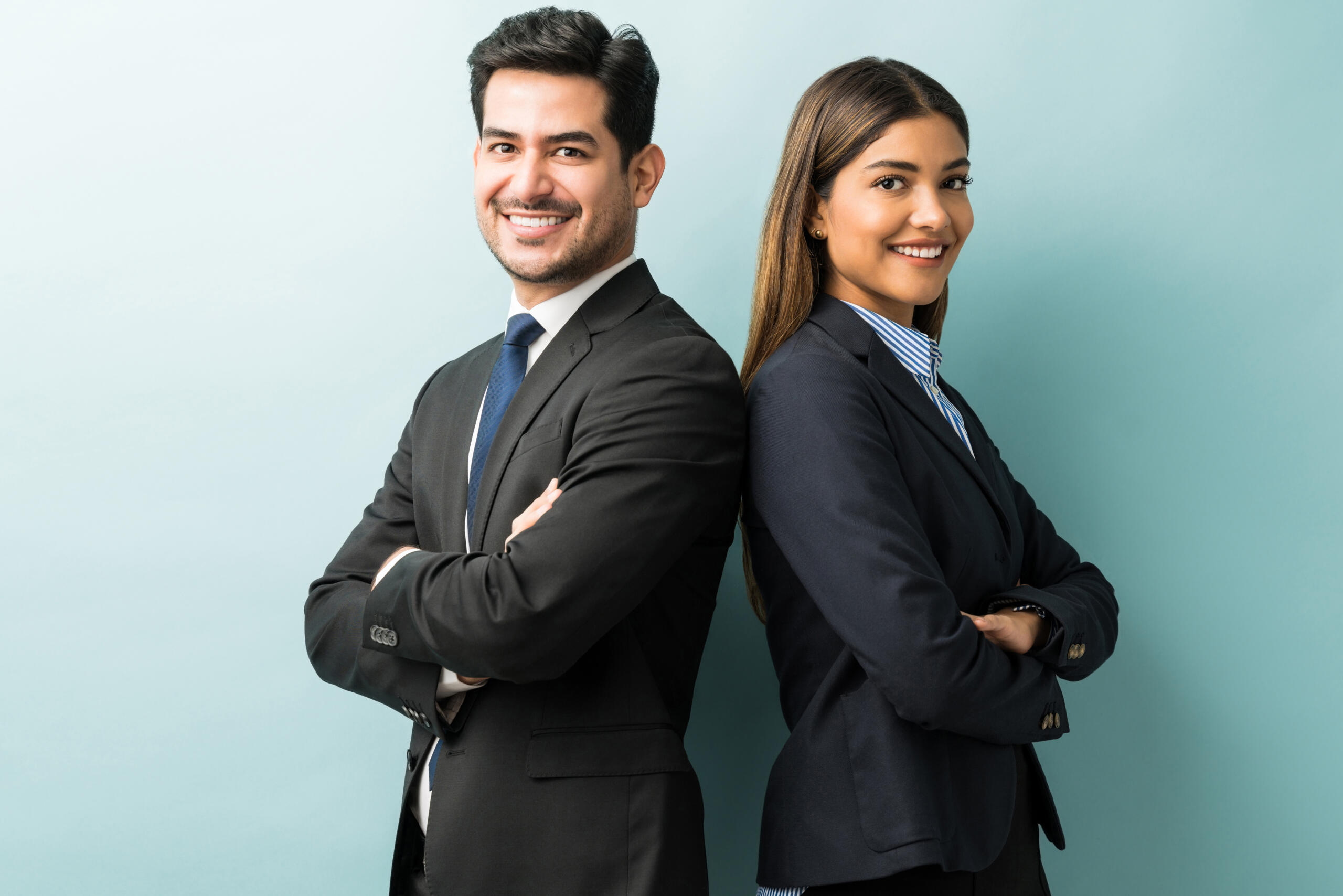 hombre y mujer vestidos con trajes formales con brazos cruzados, sonrientes y pegados espalda con espalda