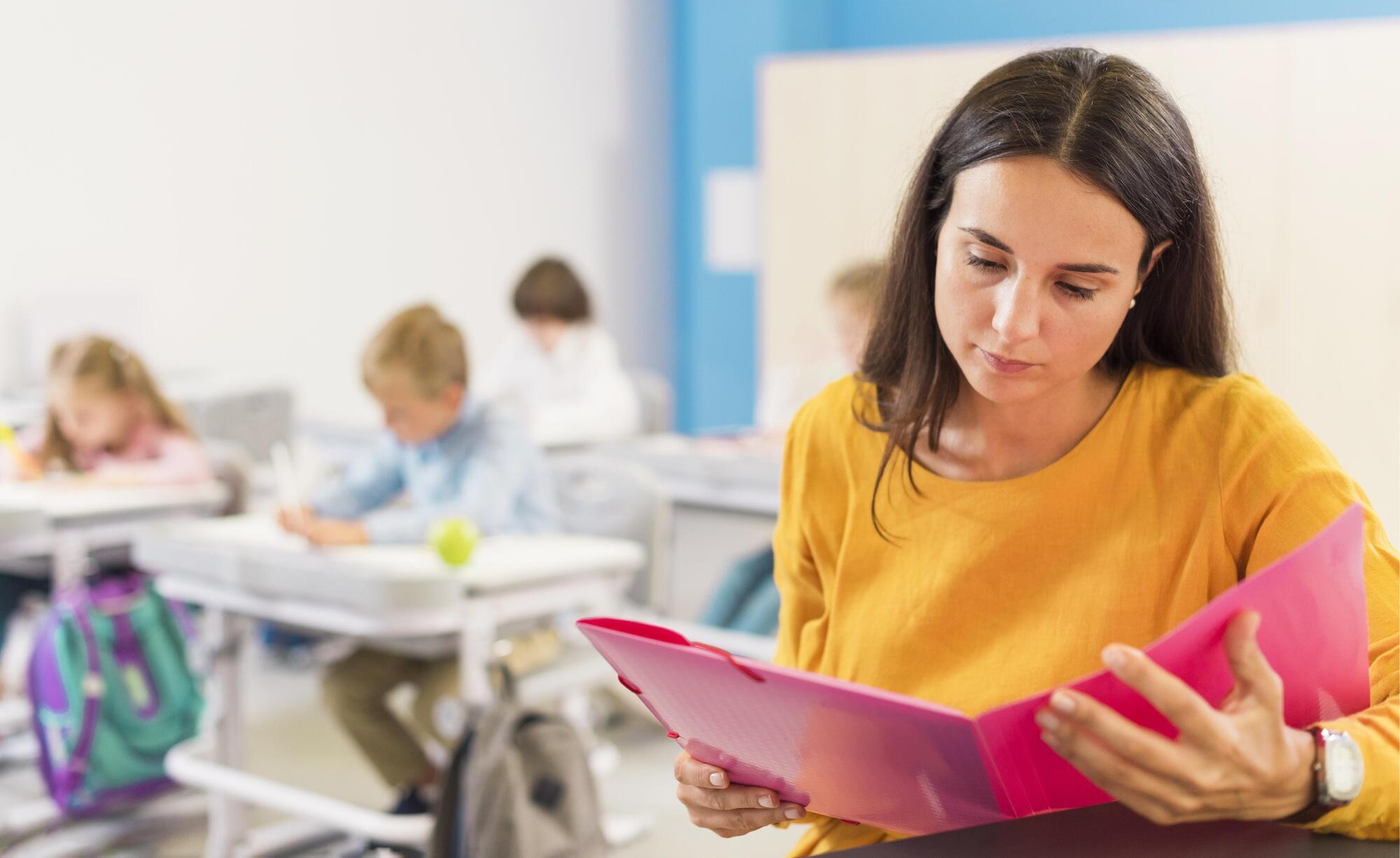 profesora en primer plano revisando un folder rosado con fondo desenfocado de niños en sus carpetas