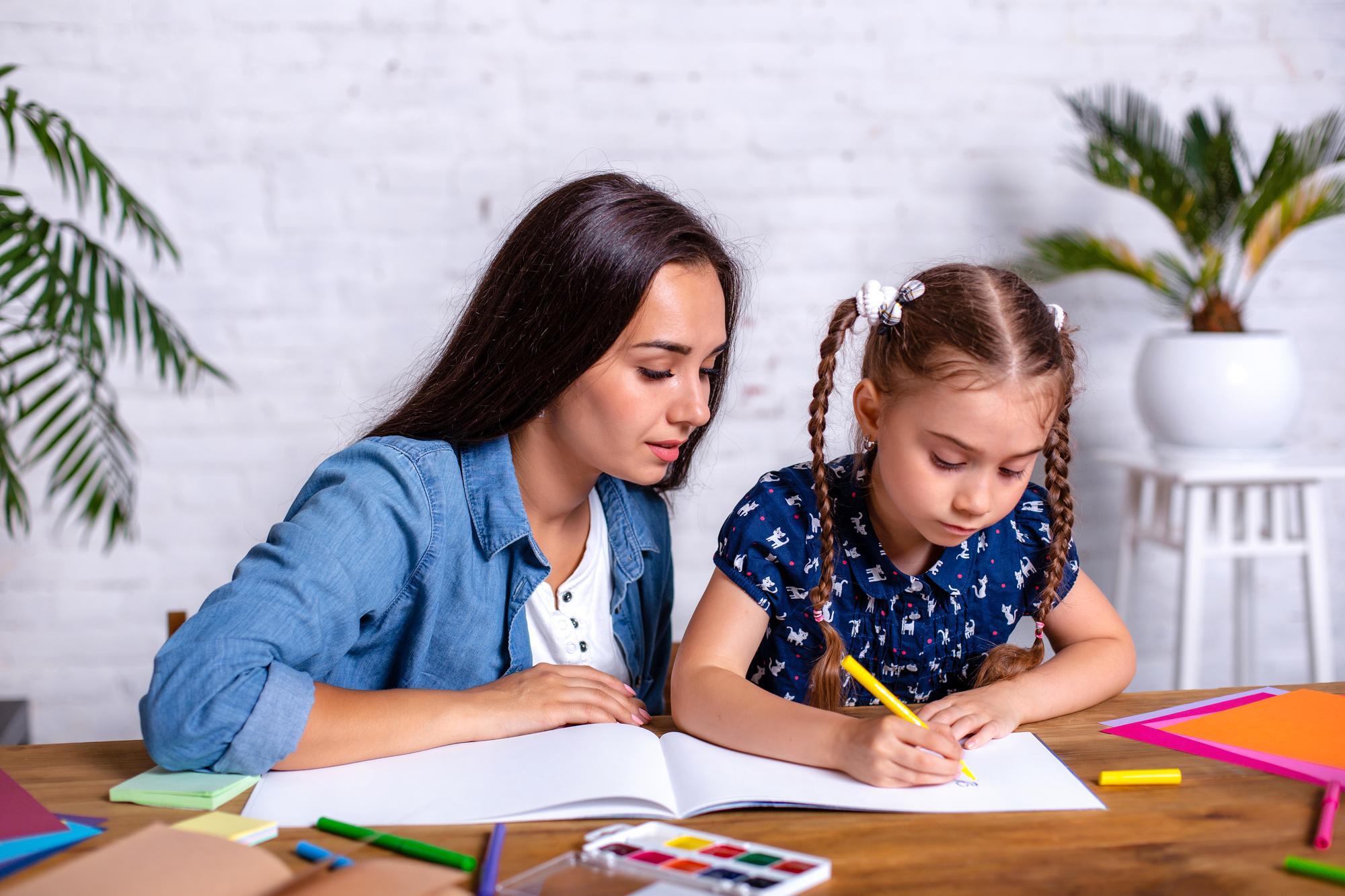 profesora enseñandole a una niña en un escritorio con cuadernos y hojas alrededor