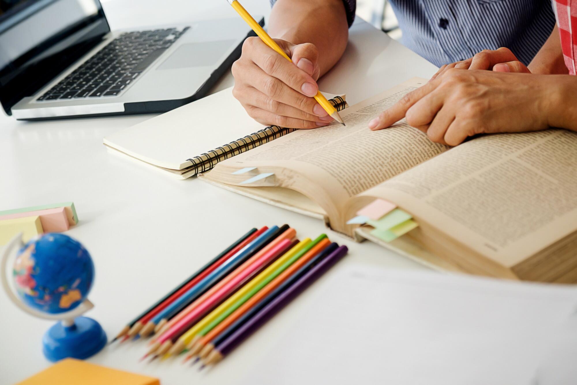 vista de un escritorio con cuadernos, lápices y manos de un profesor y alumno sobre los apuntes