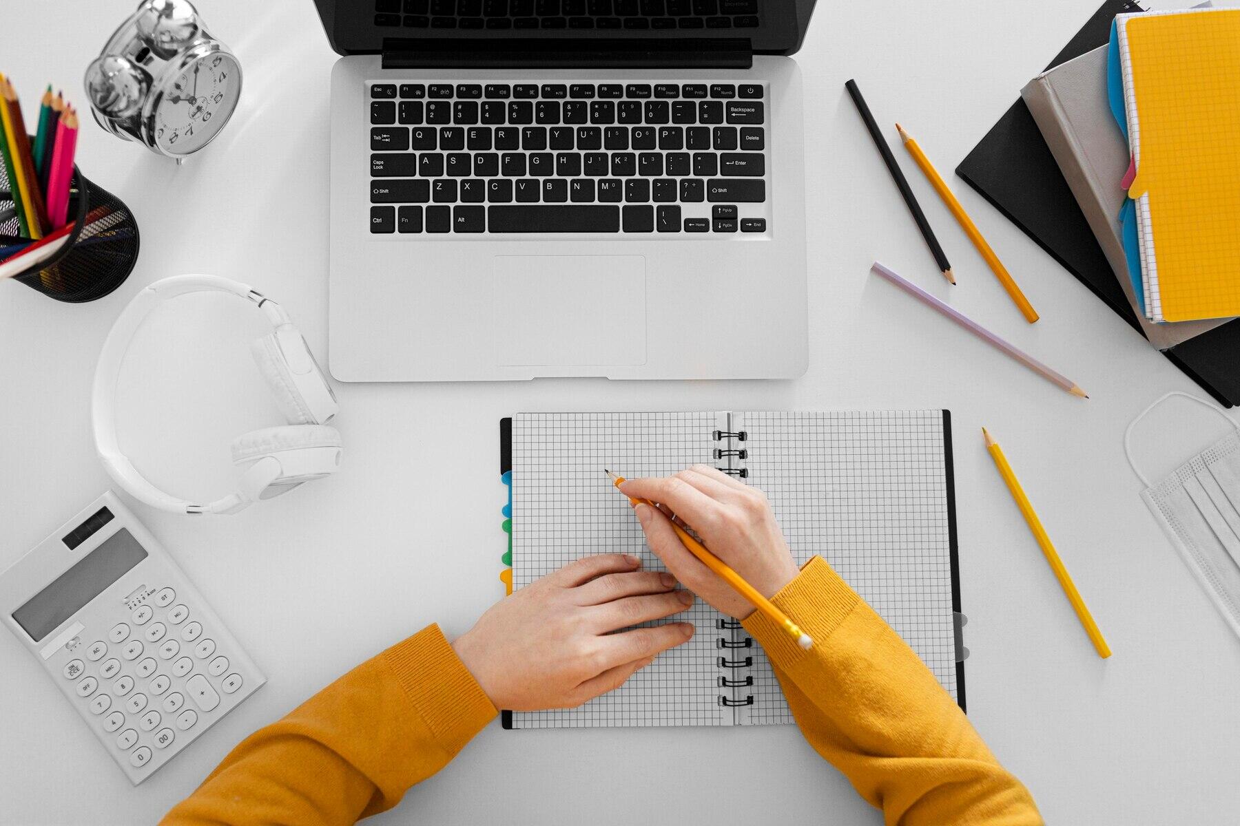 vista cenital de un ambiente de estudio, alumno con su cuaderno de notas frente a una laptop, cuadernos, calculadora y lapices alrededor