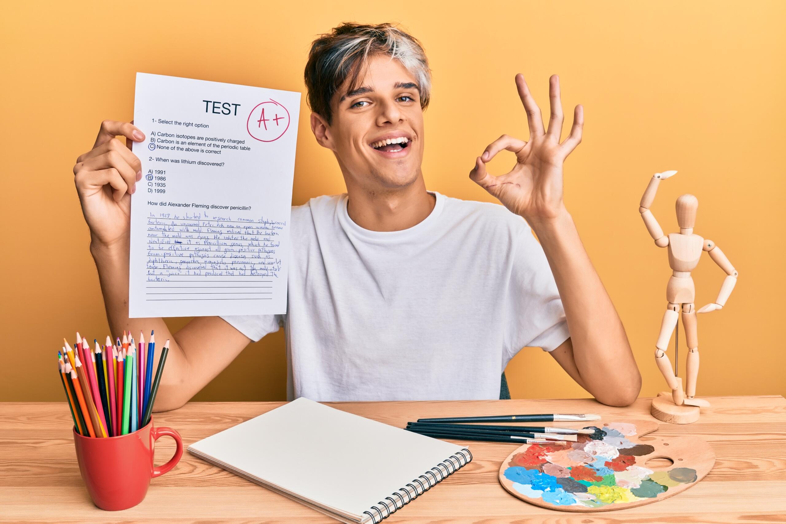 chico sonriente sosteniendo un examen A+ y haciendo un ok con la otra mano desde su escritorio lleno de anotaciones colores y hasta un maniqui de dibujo
