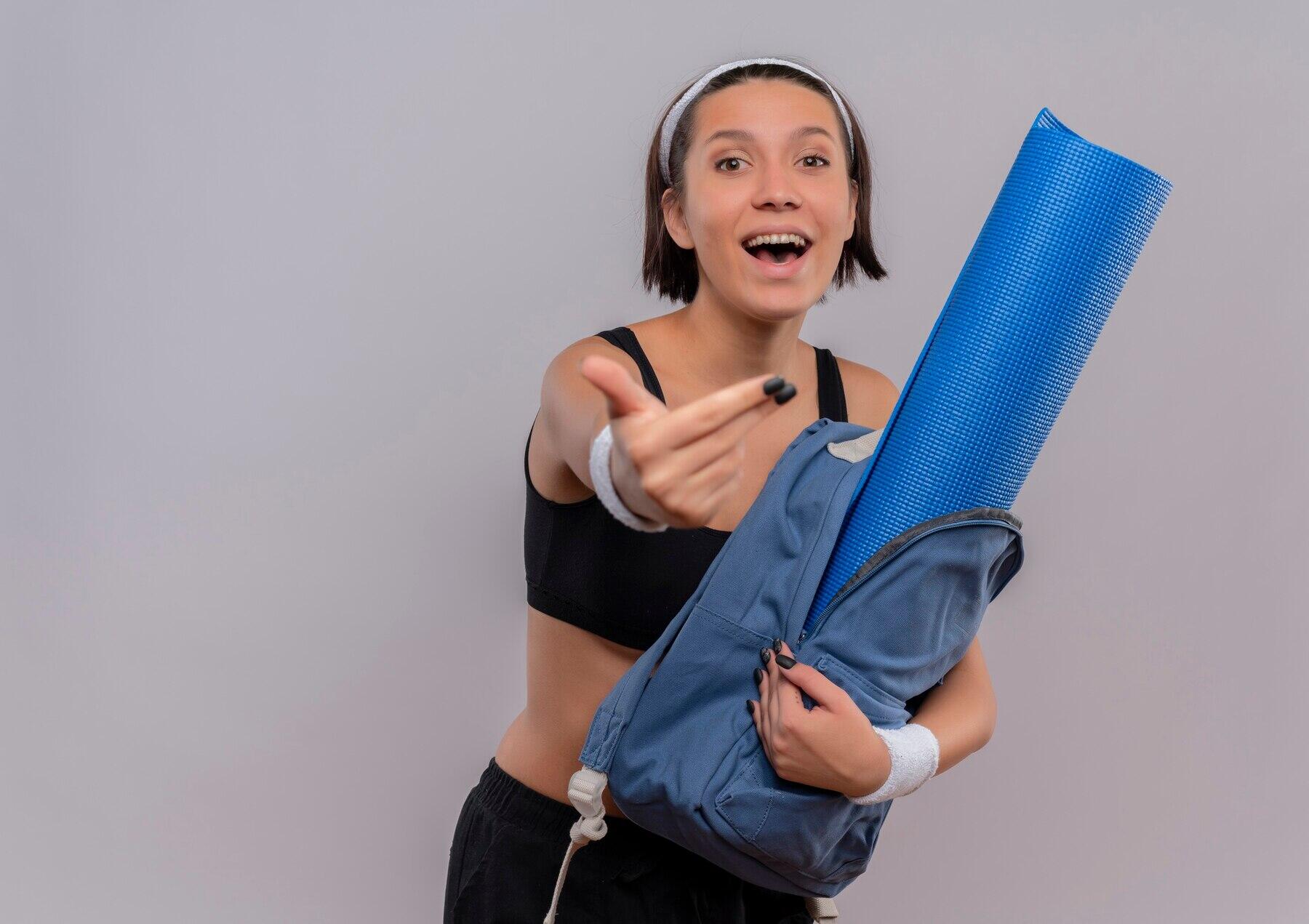 chica sonriente con ropa deportiva sosteniendo una mochila con un mat y otra mano  hacia la cámara como si llamara a alguien