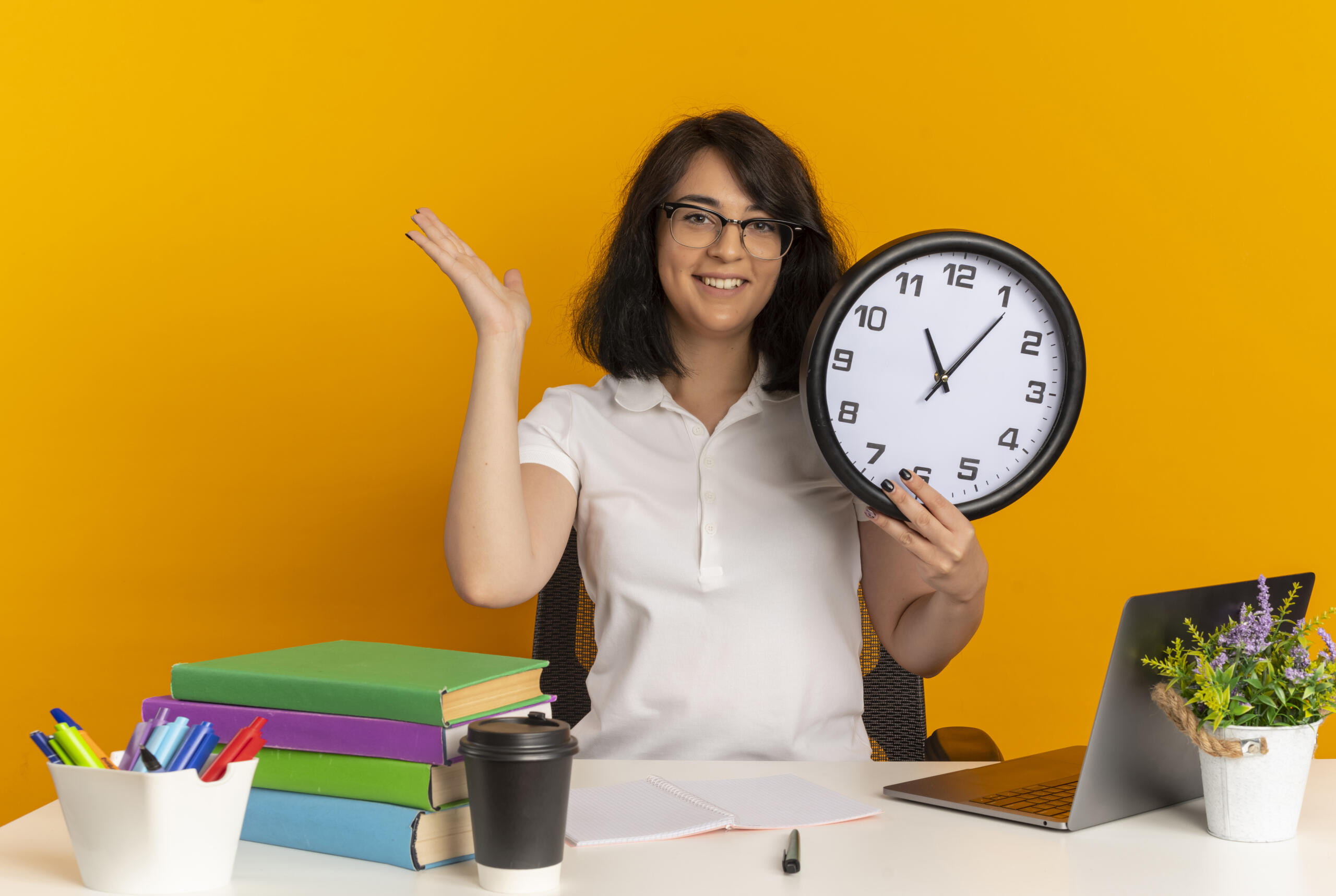 profesora sonriente sosteniendo un reloj con su escritorio frente a un fondo amarillo