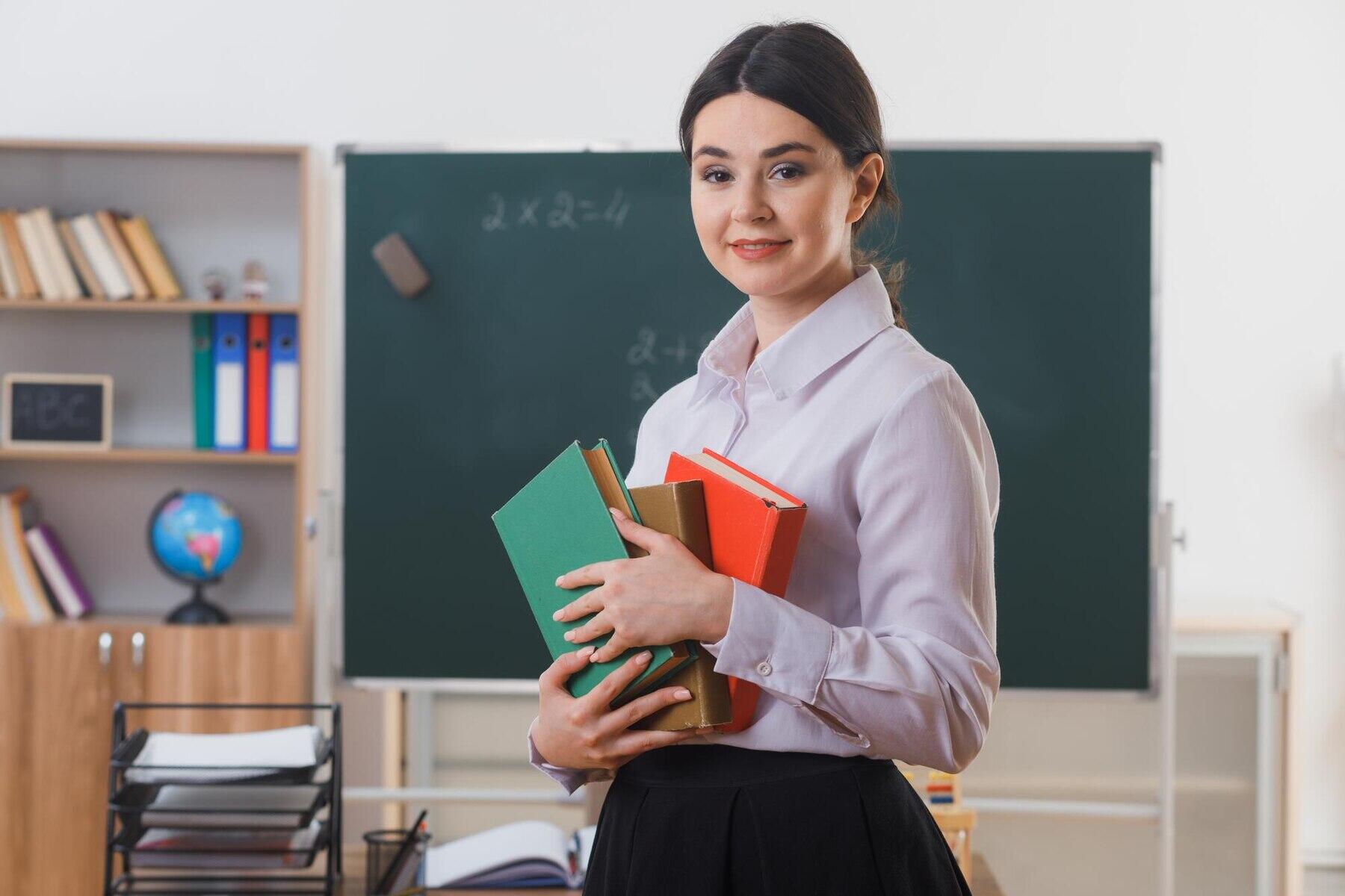 profesora sujetando libros y mirando de frente a la cámara con fondo de aula