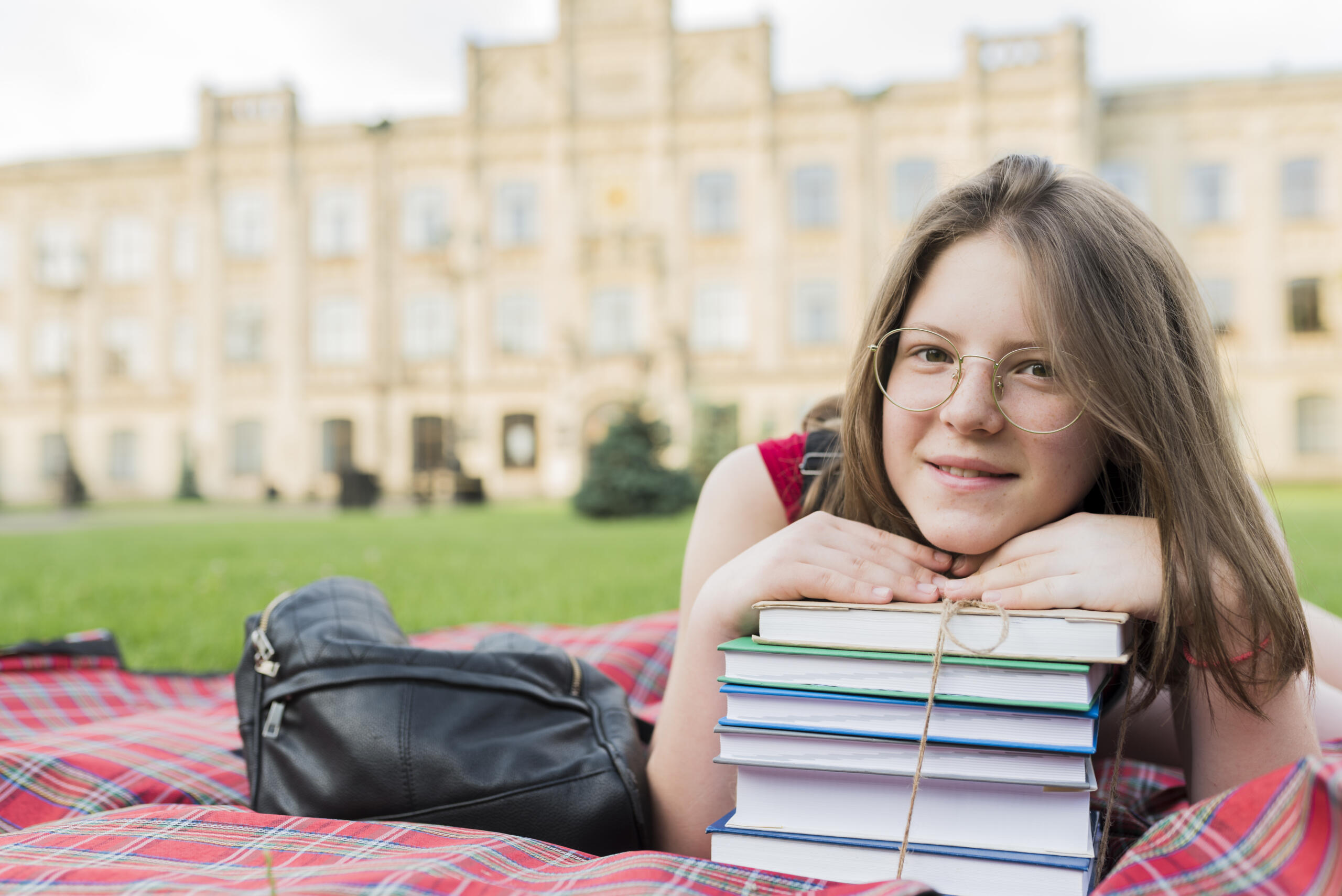 chica en un jardín frente a un colegio mirando sonriente a la cámara apoyada en libros