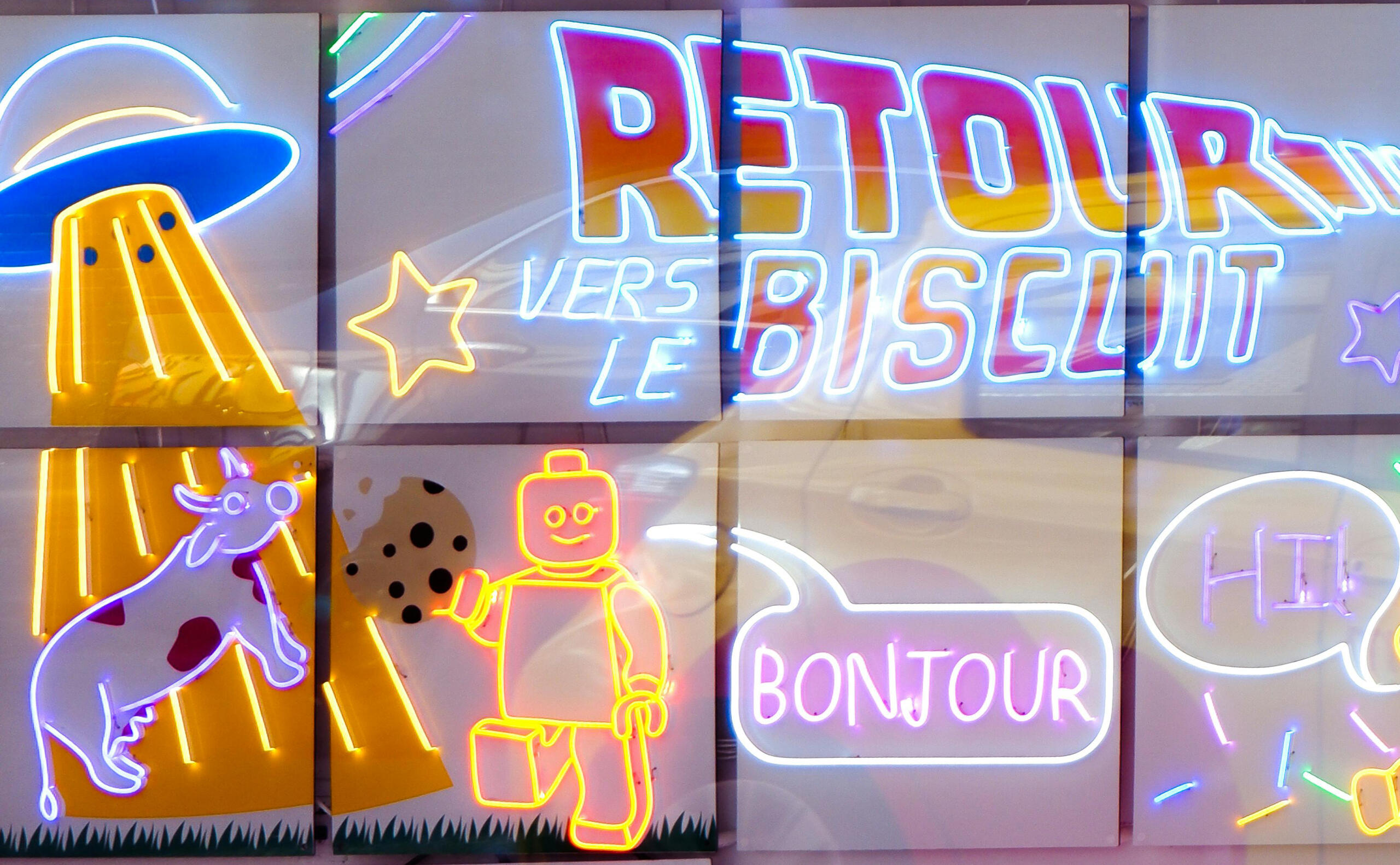 palabras en francés con luces de neón BONJOUR, RETOUR VERS LE BISCUIT junto a iconos de ovni, vaca y robot