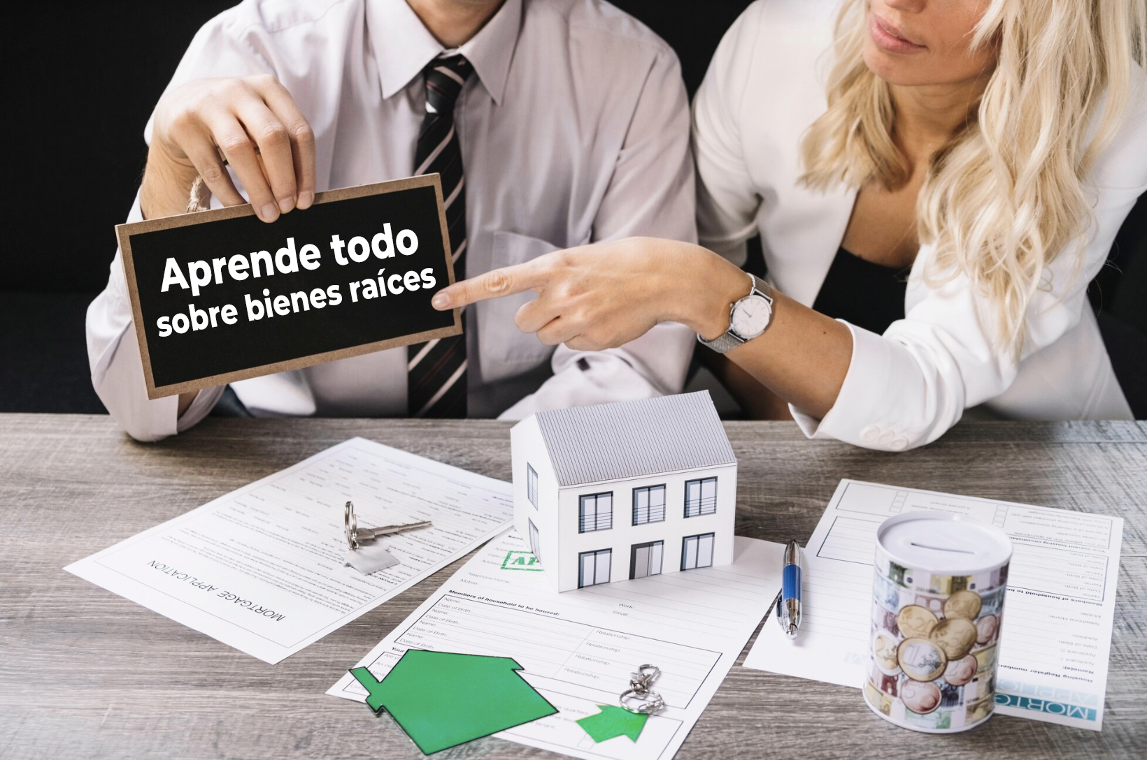 chico y chica en foto solo una parte del cuerpo mostrando un cartel que dice APRENDE TODO SOBRE BIENES RAÍCES junto a una mesa llena de papeleo de propiedad, llaves, monedas y una maqueta de casa