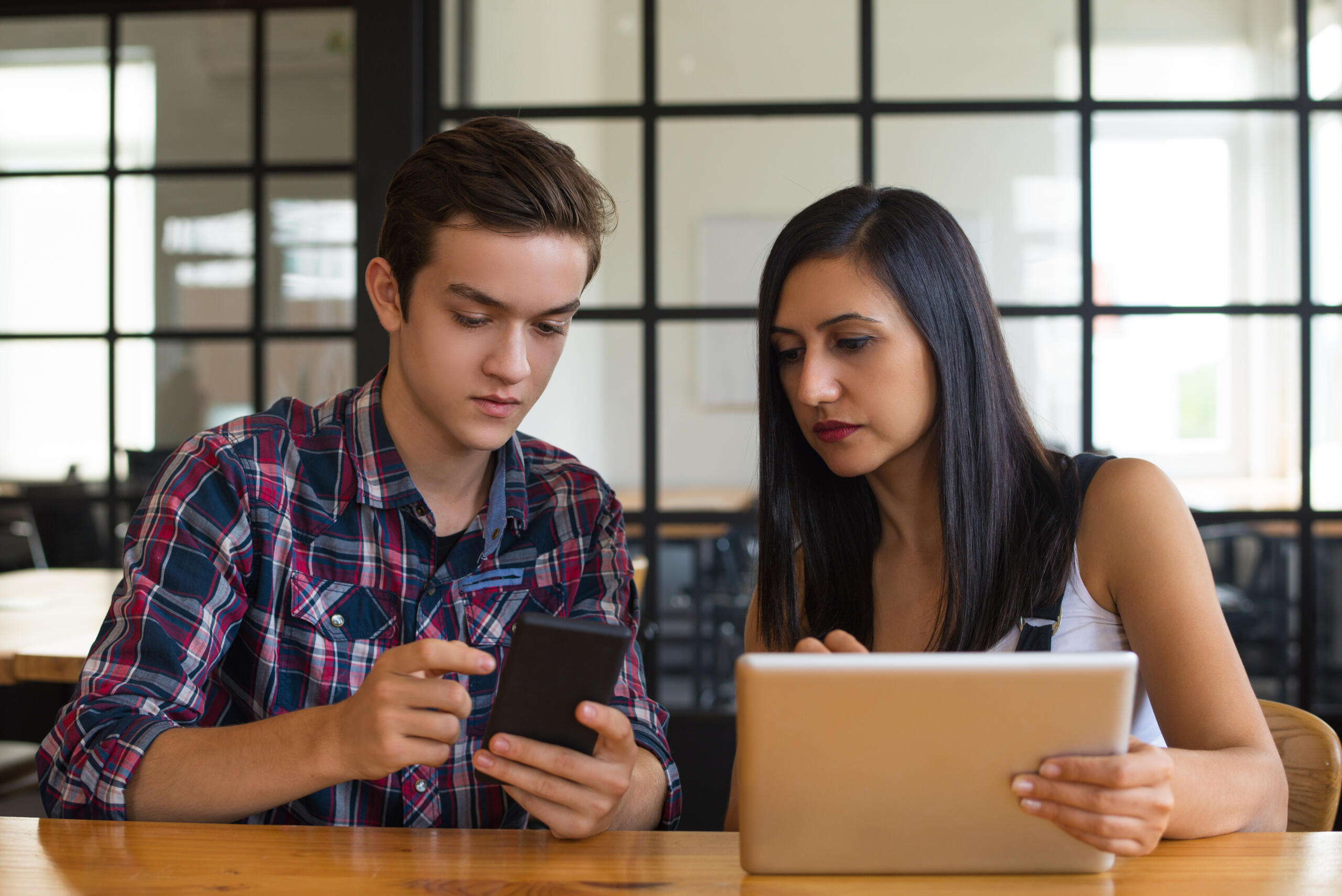 joven mostrándole algo en su celular a la maestra que sostiene un ipad
