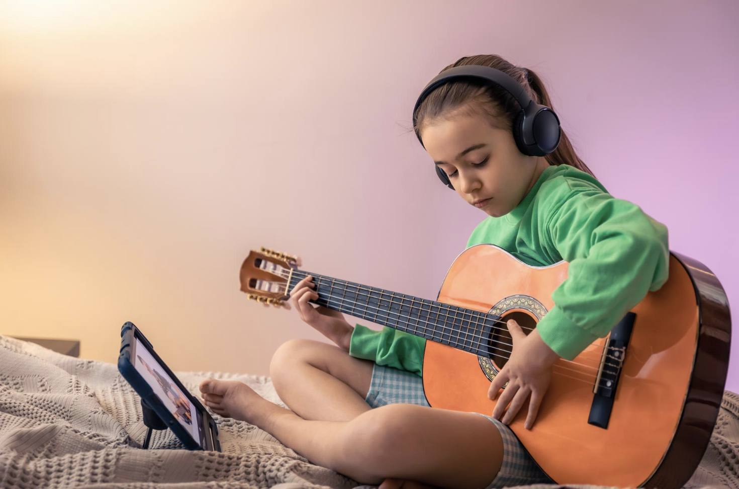 niña en una habitacion de tonos pasteles tocando la guitarra frente a su tablet con audifonos
