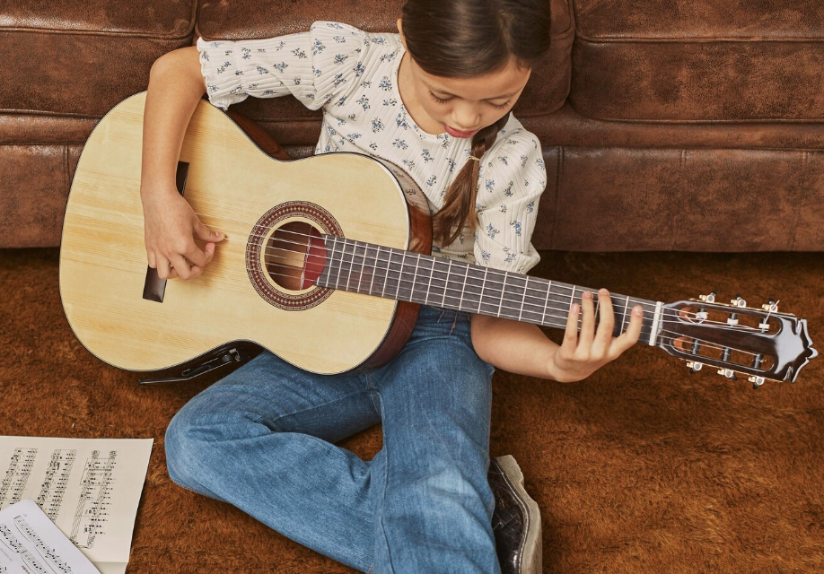 niña sentada en el suelo alfombrado tocando una guitarra junto a unas partituras abiertas junto a ella