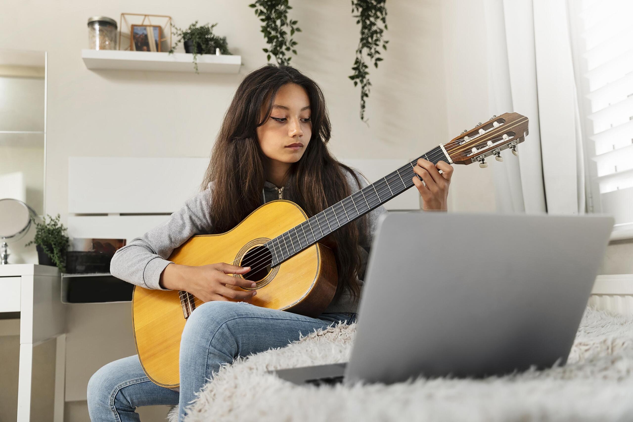 niña sentada en un mueble tocando la guitarra frente a su laptop