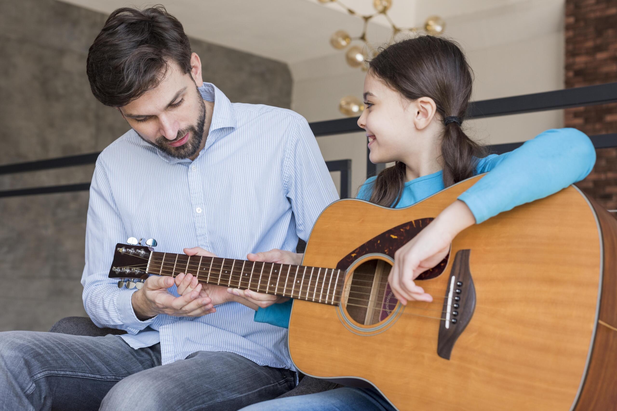niña sonriente con una guitarra en manos junto al profesor que está enseñándole a tocar guitarra