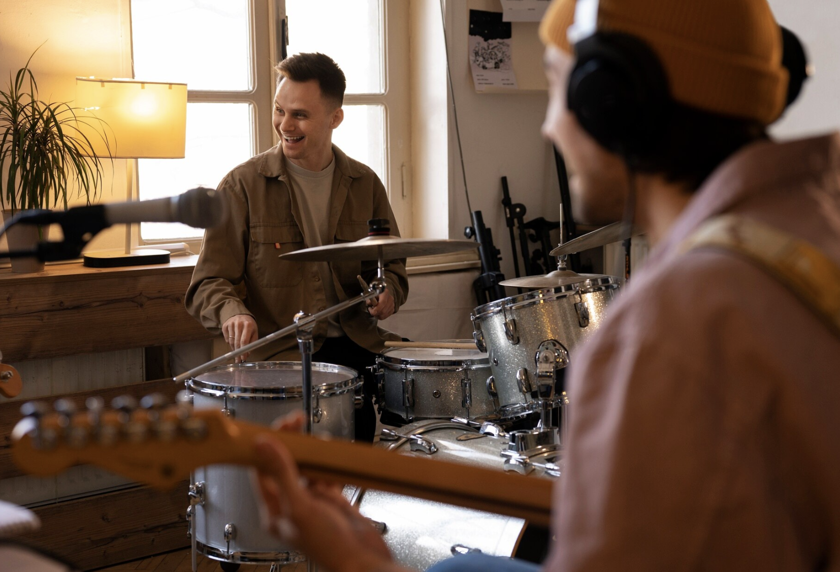 chico sonriente tocando la bateria teniendo en un primer plano desenfocado a otro chico tocando la guitarra