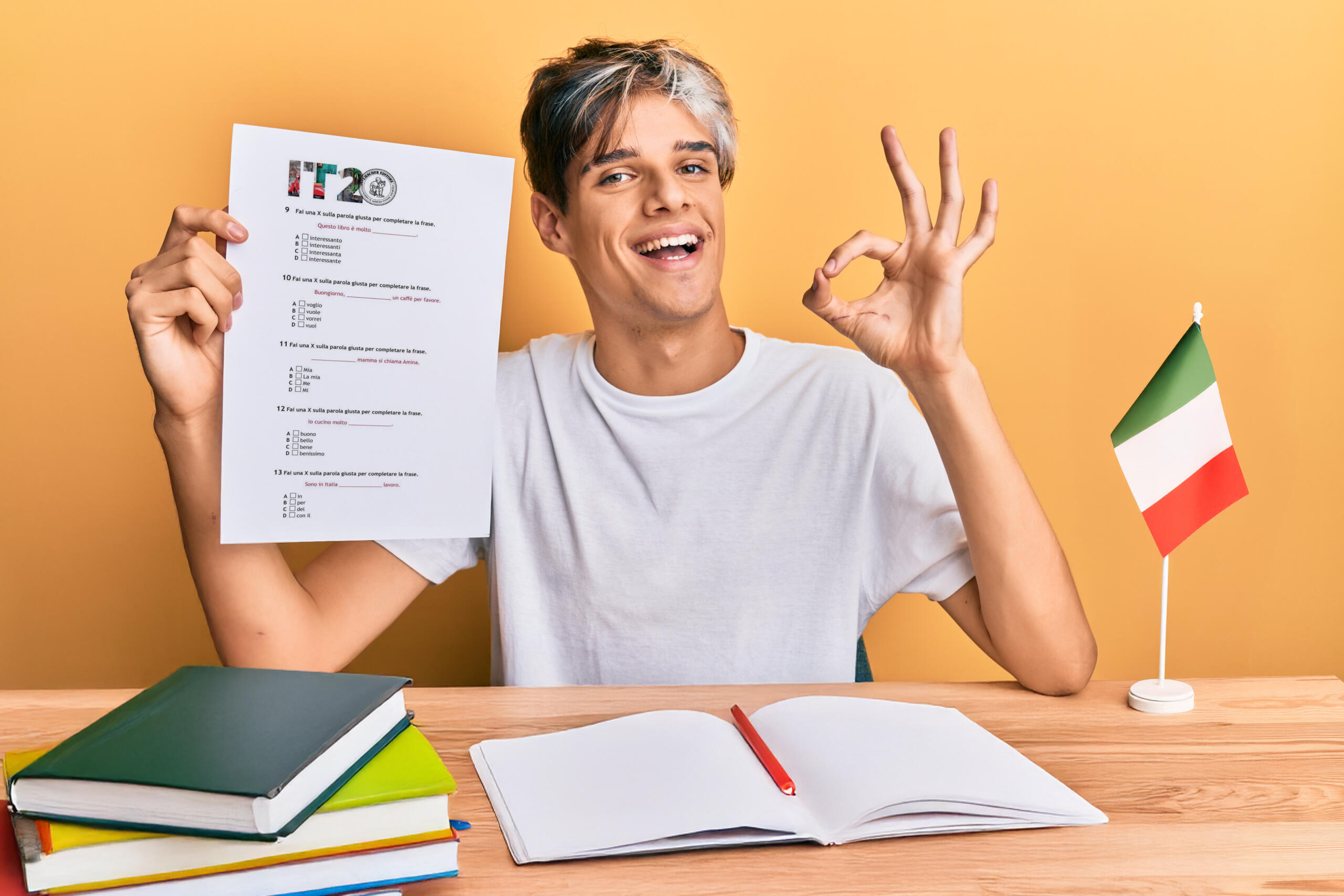 chico sonriente en su escritorio mostrando seña ok con una mano y sosteniendo una prueba de italiano en la otra