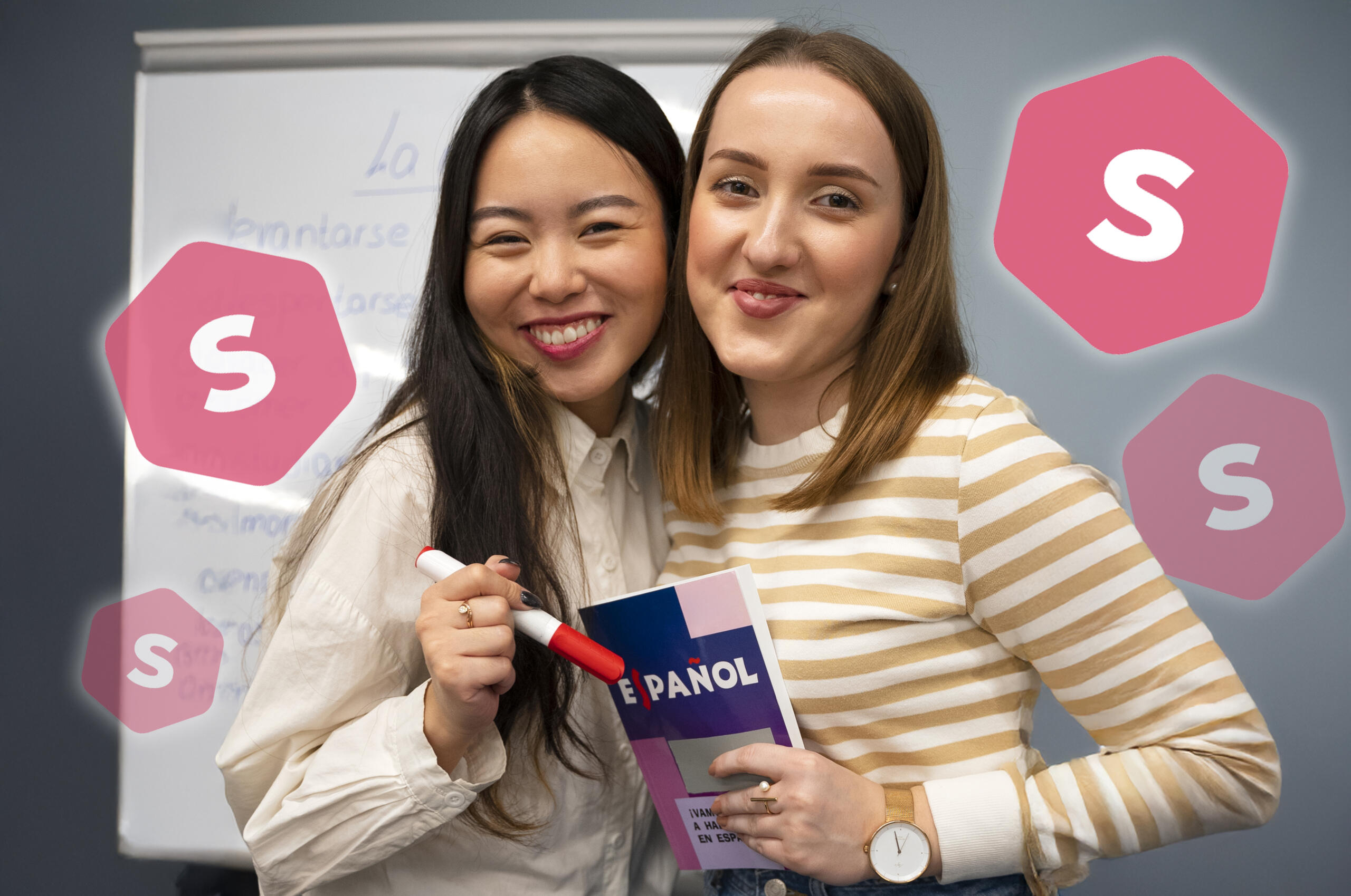 dos chicas sonrientes mirando a la cámara con un libro de español y fondo de pizarra con palabras escritas en español