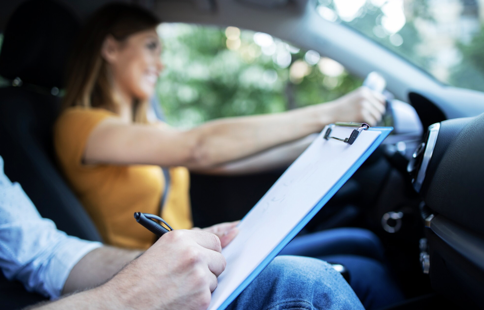 primer plano de manos de instructor haciendo anotaciones con vista de alumna desenfocada en asiento de conductor