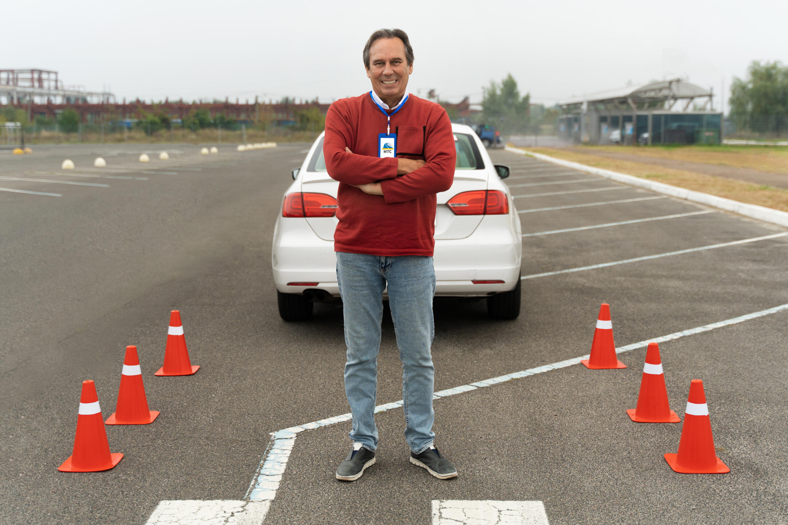 instructor de manejo cruzado de brazos sonriente mirando a la cámara con auto y circuito de manejo de fondo
