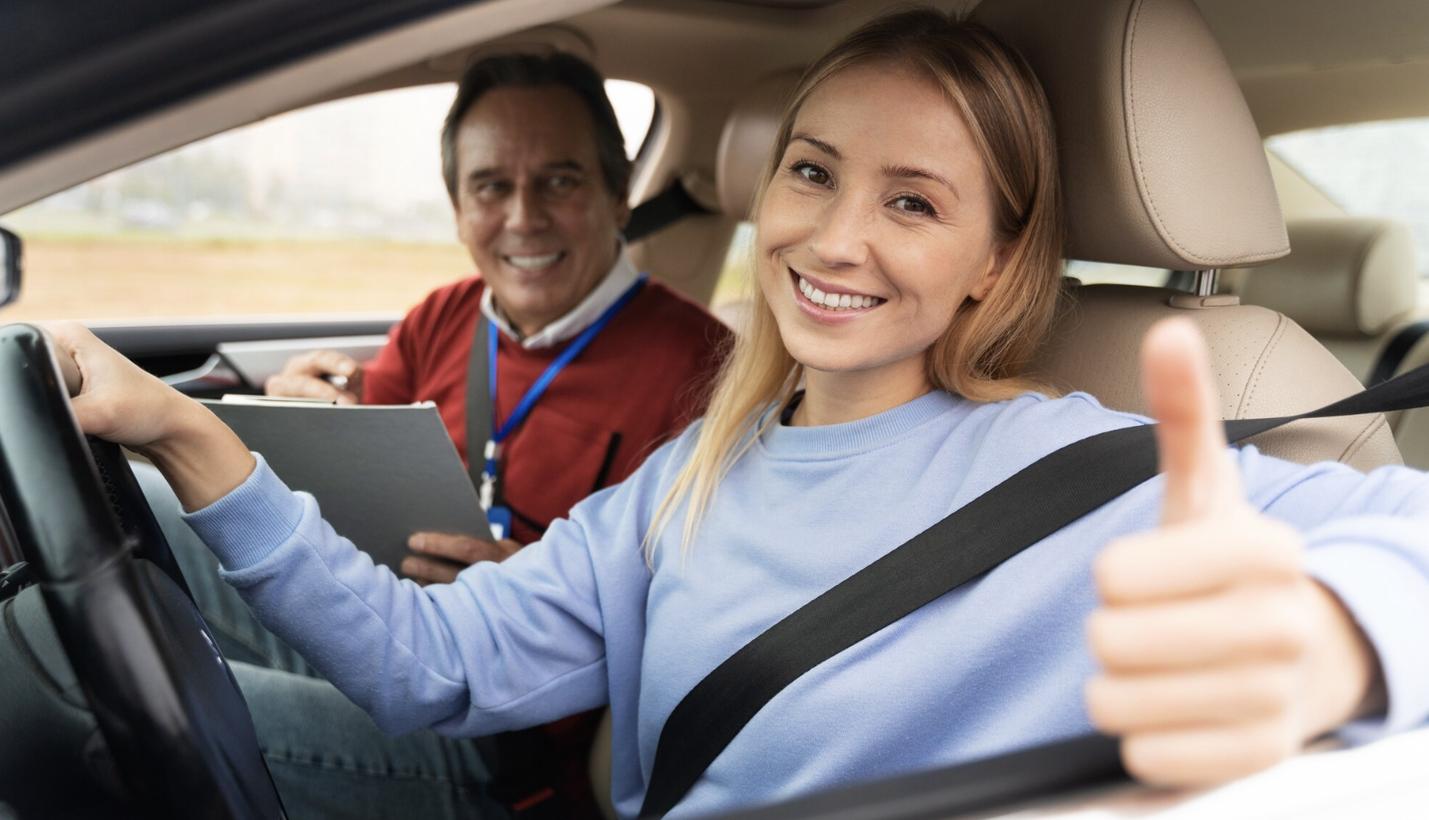 chica sonriente al volante con mano haciendo señal de ok con su instructor de fondo como copiloto