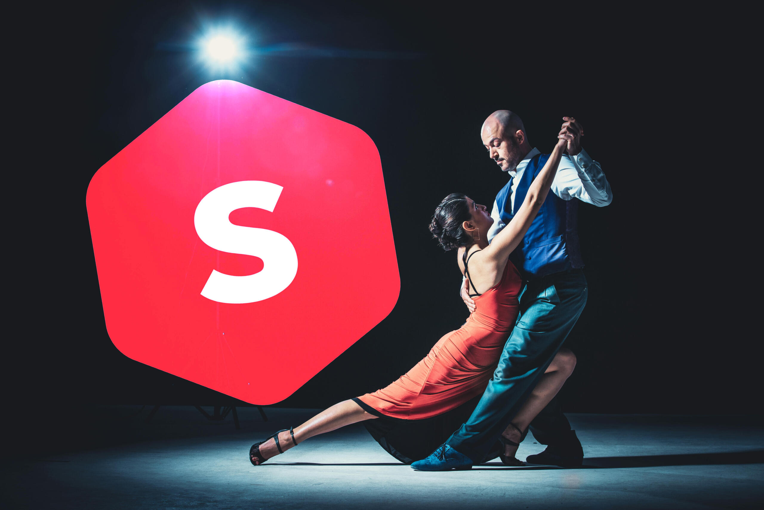 Pareja bailando tango en un escenario a oscuras con un reflector iluminándolos junto al logo de Superprof