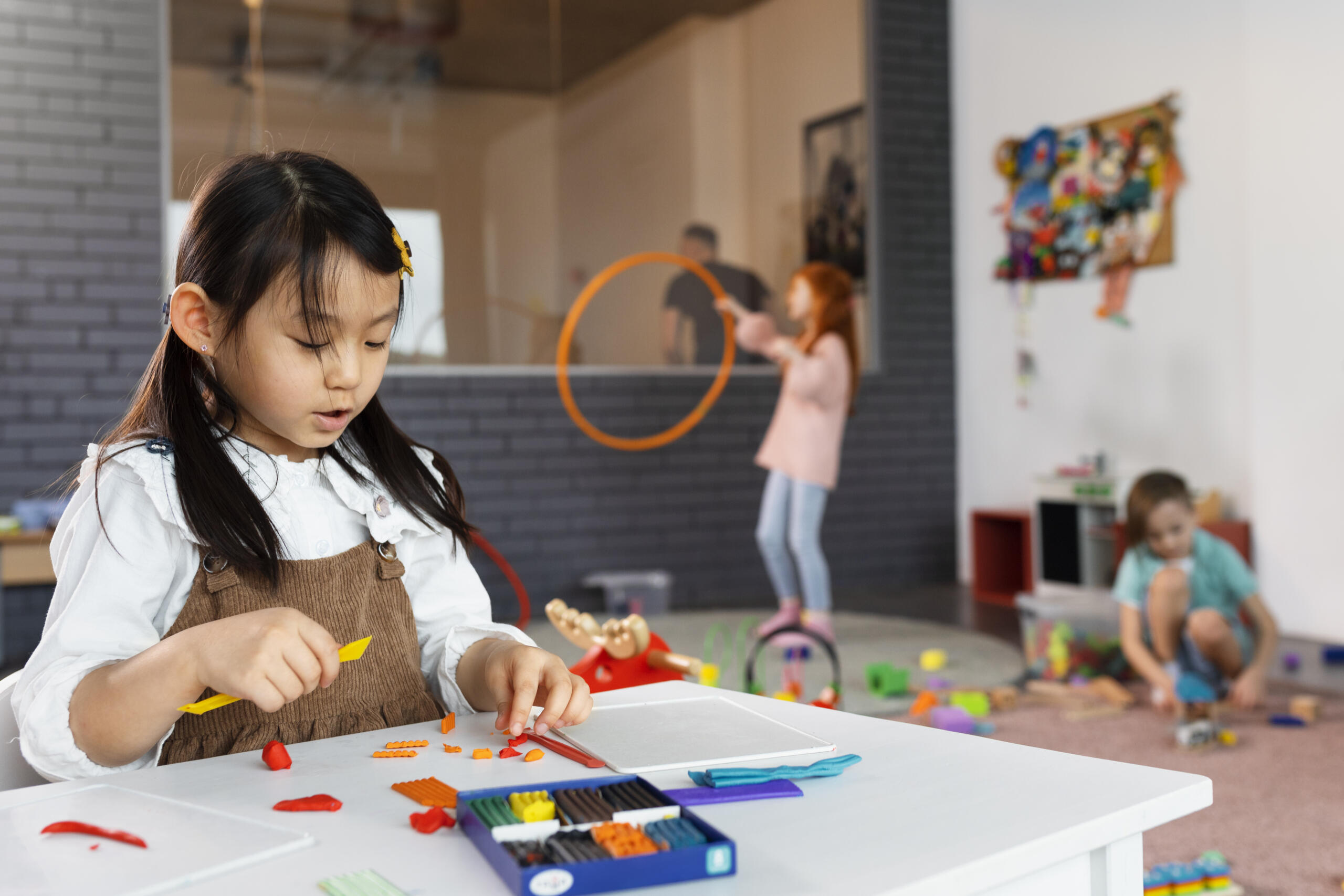 niña jugando con plastilina en una mesa y de fondo más niños haciendo otras actividades