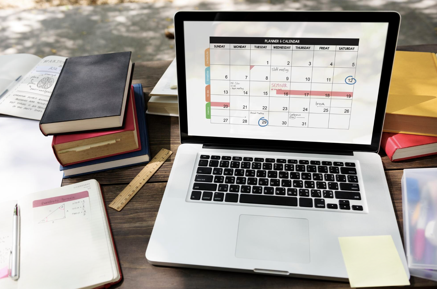 laptop con un calendario en pantalla rodeada de libros y cuaderno de apuntes por todos lados.