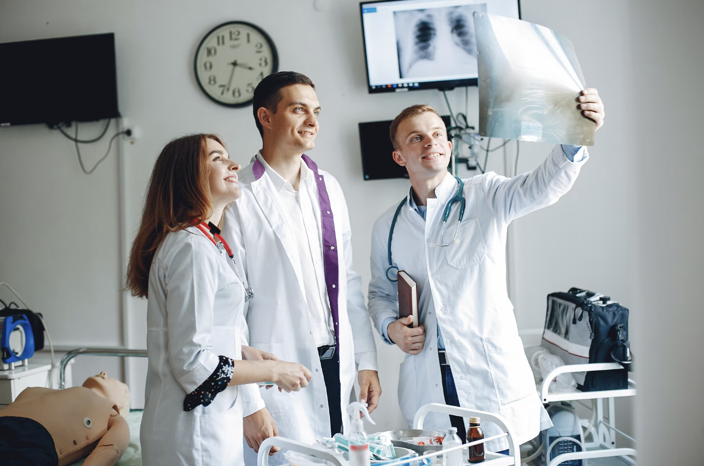 grupo de tres estudiantes de medicina viendo una radiografía y en el fondo se ve la sala de práctica