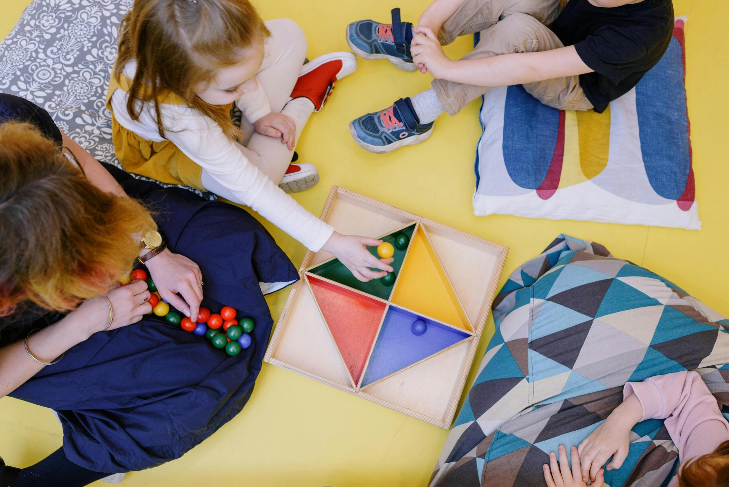 toma cenital de maestra sentada en el suelo con tres niños haciendo una actividad montessori con una caja con triangulos de colores y cuentas coloridas de madera