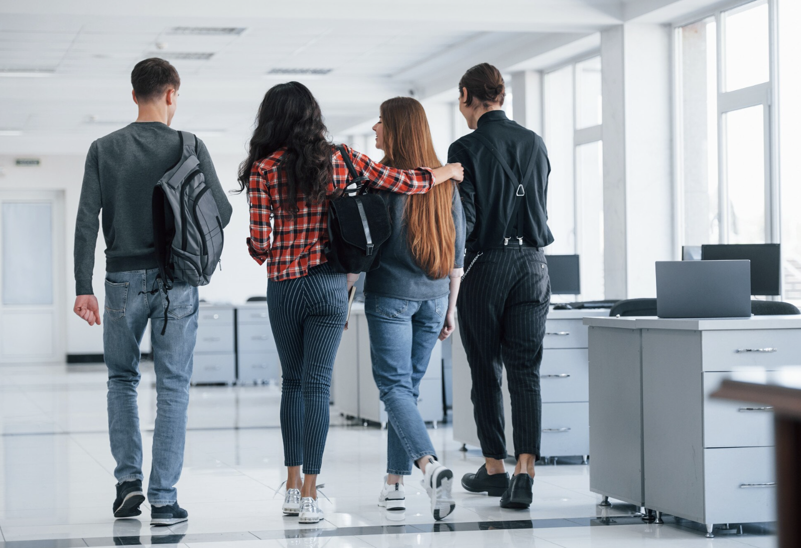 se ve de espaldas a un grupo de cuatro estudiantes hombres y mujeres caminando en una sala de estudio juntos