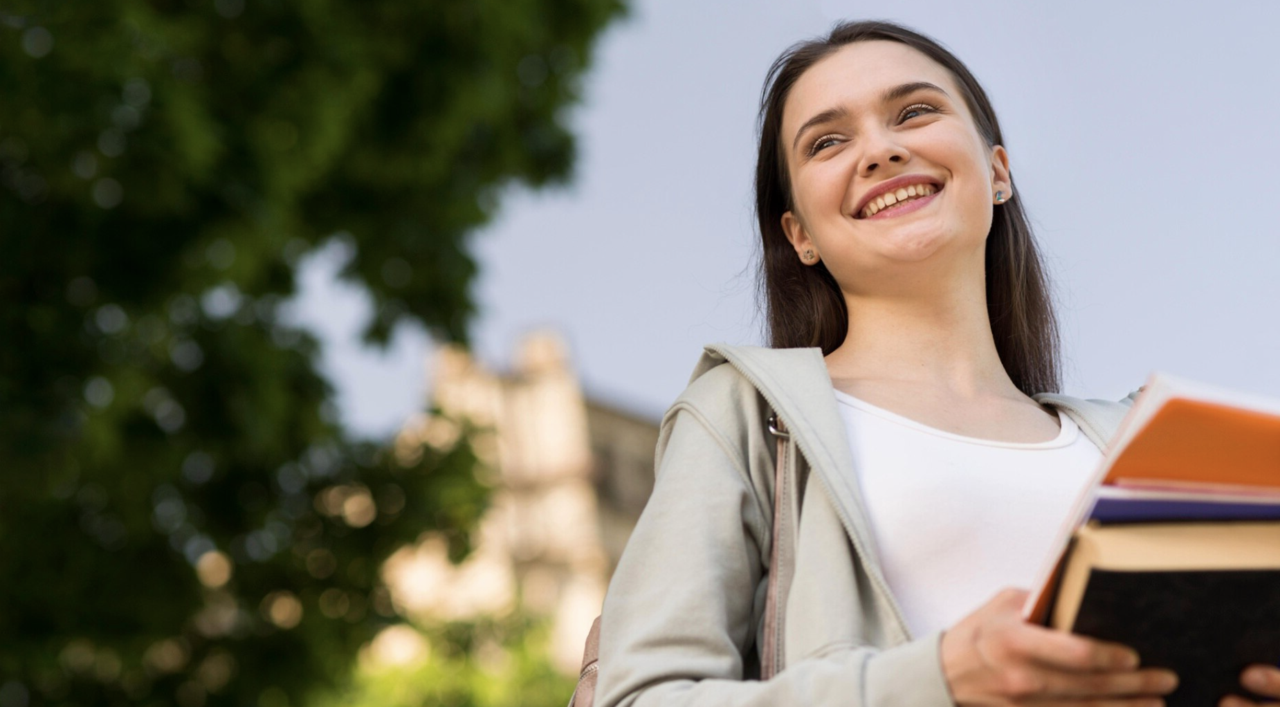 chica sonriente sosteniendo libros con un arbol de fondo