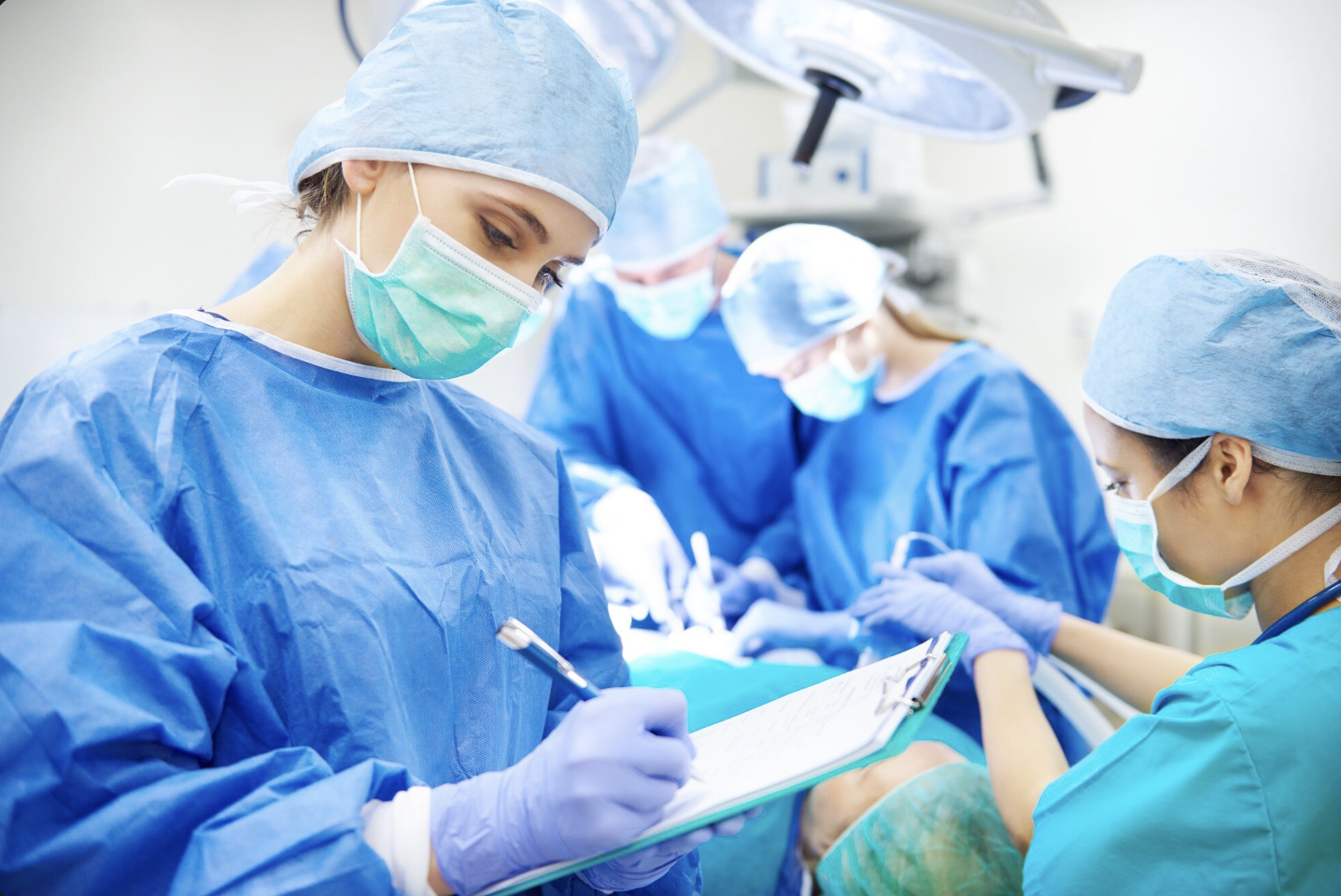 doctora realizando anotaciones con uniforme de cirugia y de fondo se ve a un grupo de dos médicos y una enfermera en medio de una operación