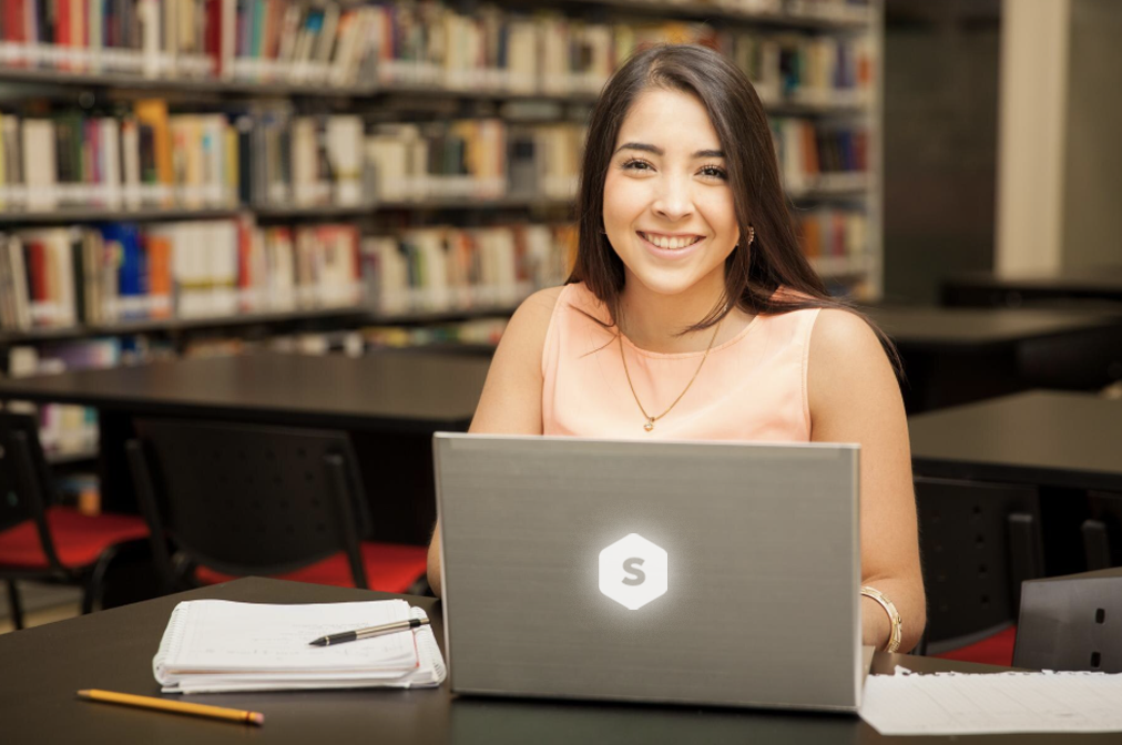 chica sonriente en escritorio de biblioteca mirando a la cámara. con laptop que tiene logo de superprof en la tapa