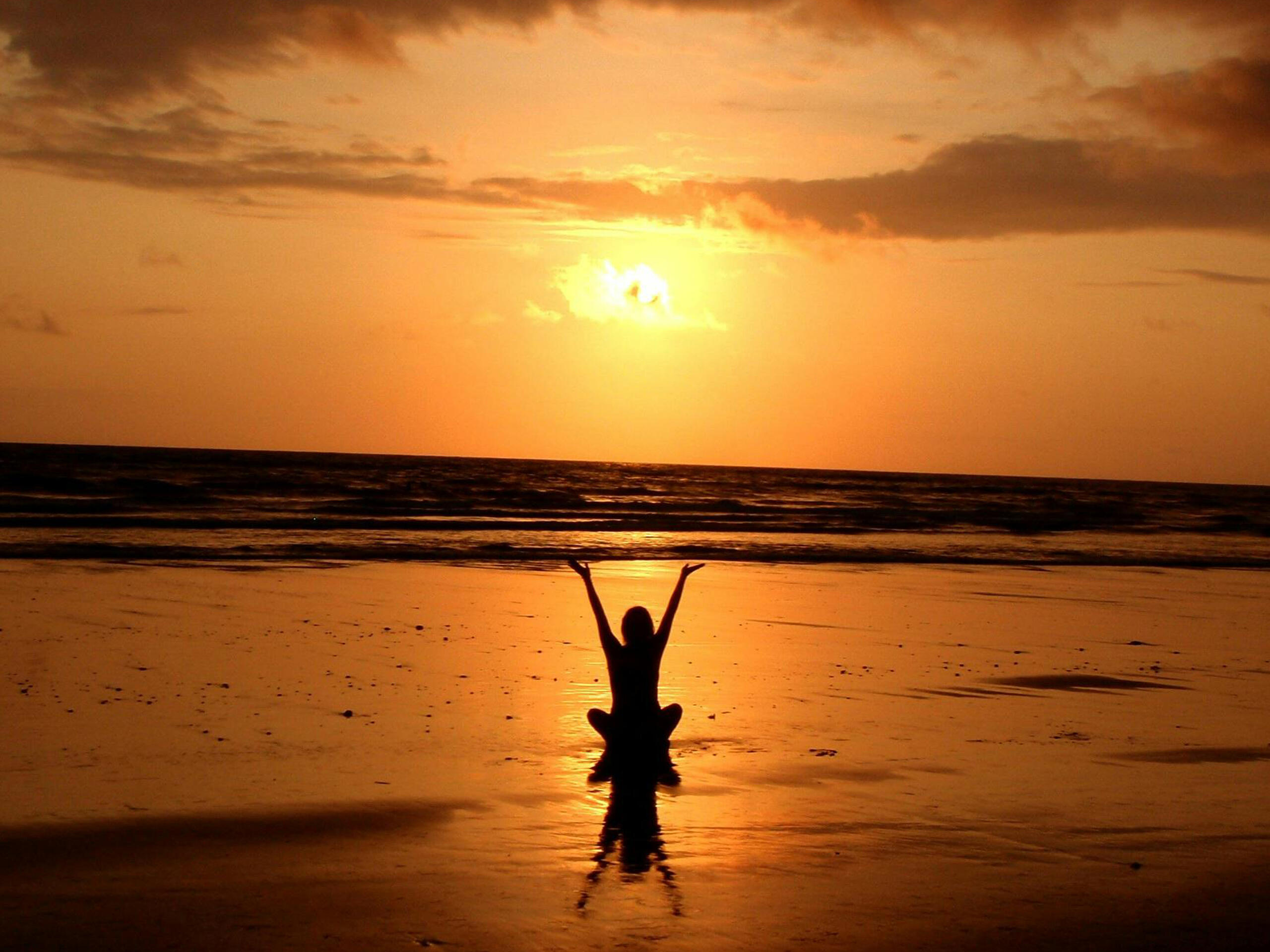 silueta de chica sentada en la arena húmeda frente al mar con los brazos extendidos con el sunset