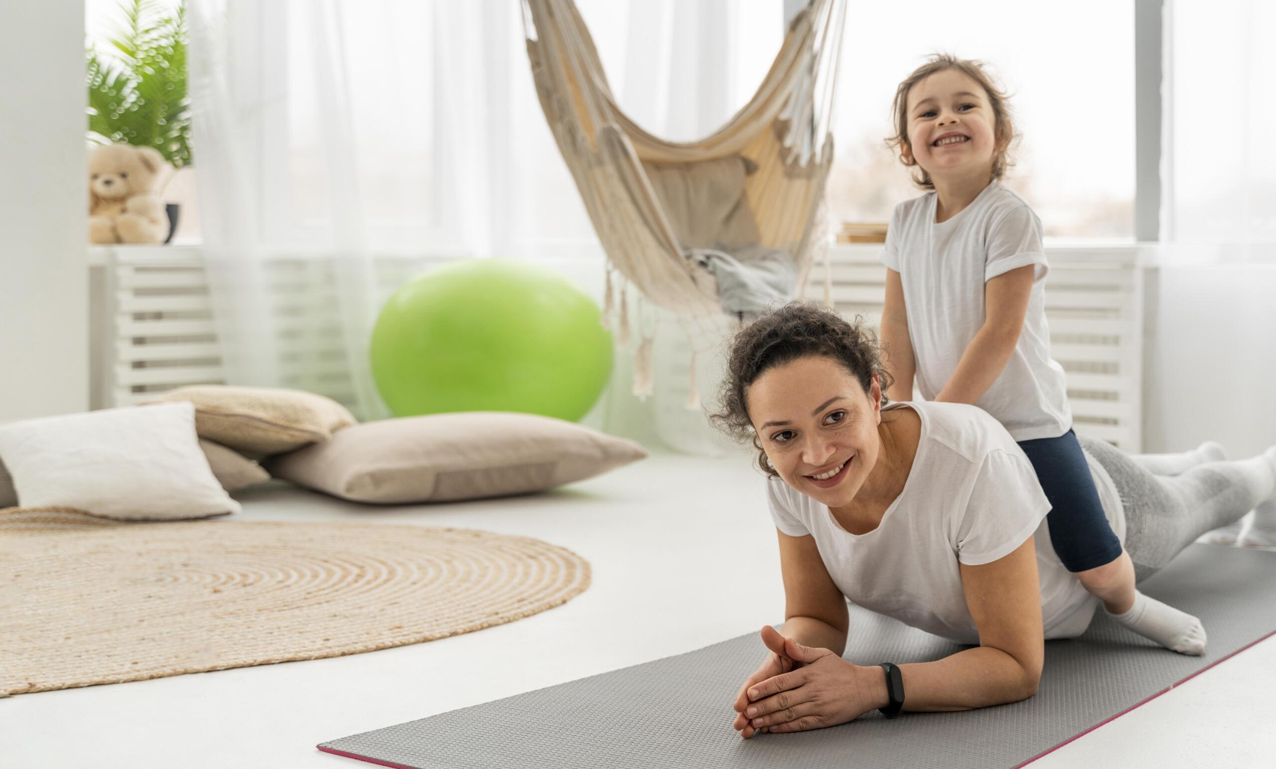 Señora haciendo un plank en la sala de su casa con una niña encima de su espalda sonriente