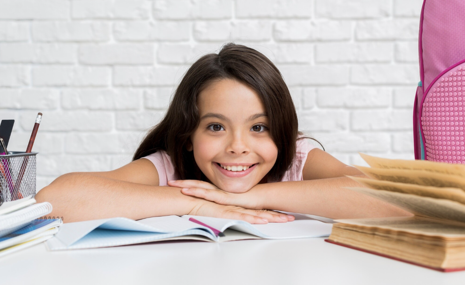 niña sonriente mirando a la cámara apoyada sobre libro abierto en su escritorio