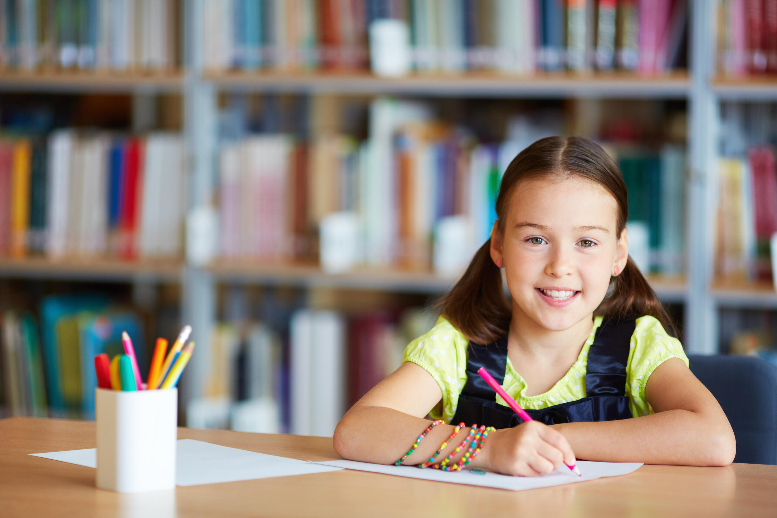 niña sonriente en una mesa escribiendo con lapices de colores y con un fondo de librero
