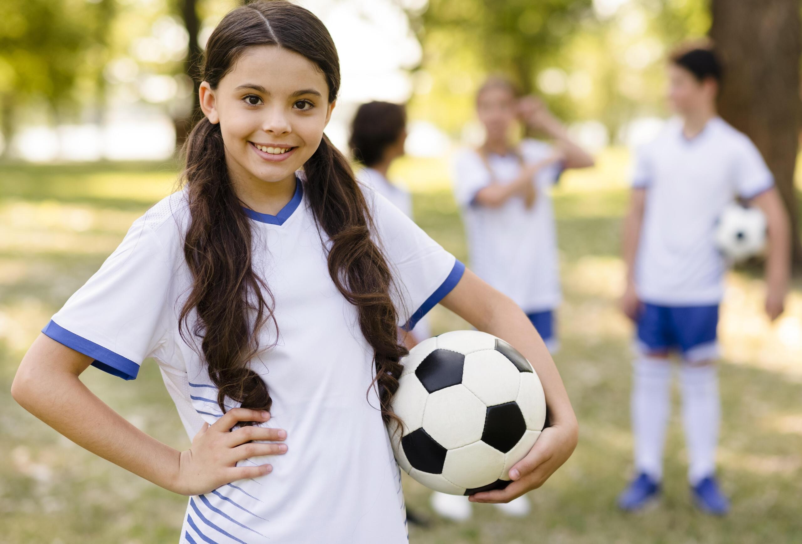 niña en primer plano con un uniforme y pelota de futbol sonriente a la cámara con un grupo de niños de fondo