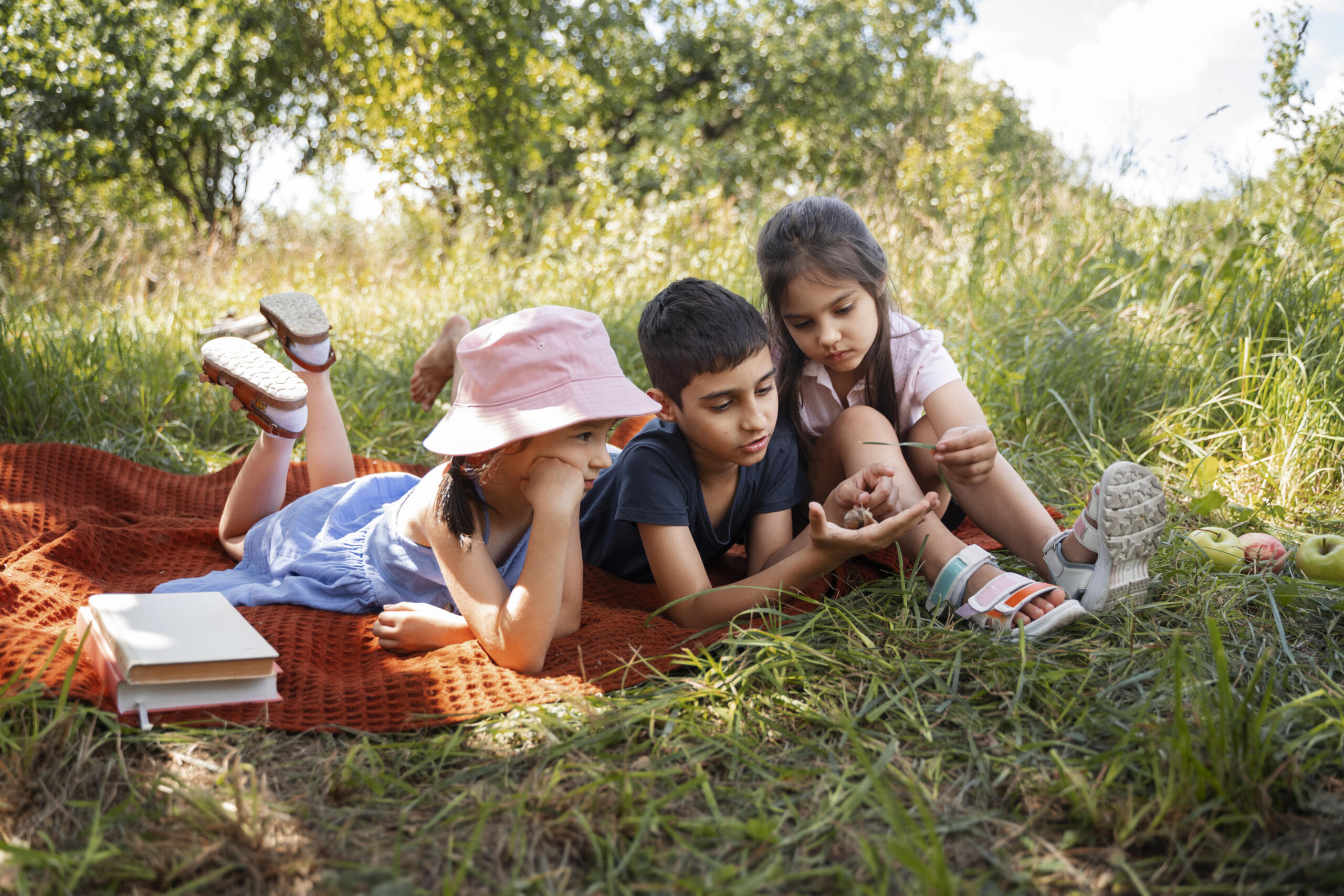 tres niños echados boca abajo jugando con las hojas en un manto en el césped junto a libros