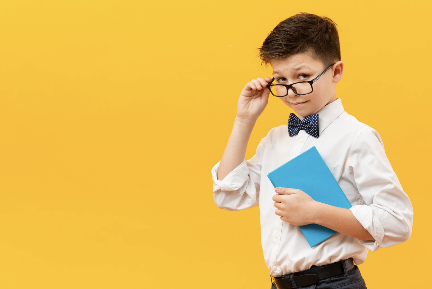 niño con michi y traje sosteniendo unos libro y sujetando unos lentes en el puente de la nariz mientras mira la camara con un fondo amarillo