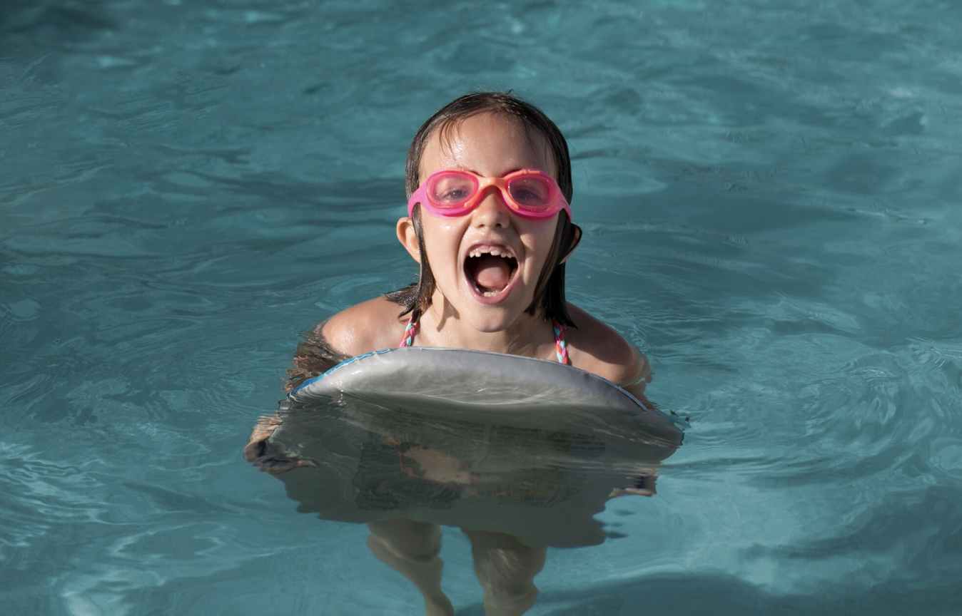 niña sonriente en medio de la piscina con lentes y tabla de natación