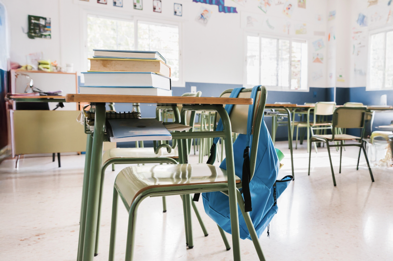 carpeta de escuela con libros apiladores y mochila colgada en el respaldar de la silla con ambiente de aula de fondo