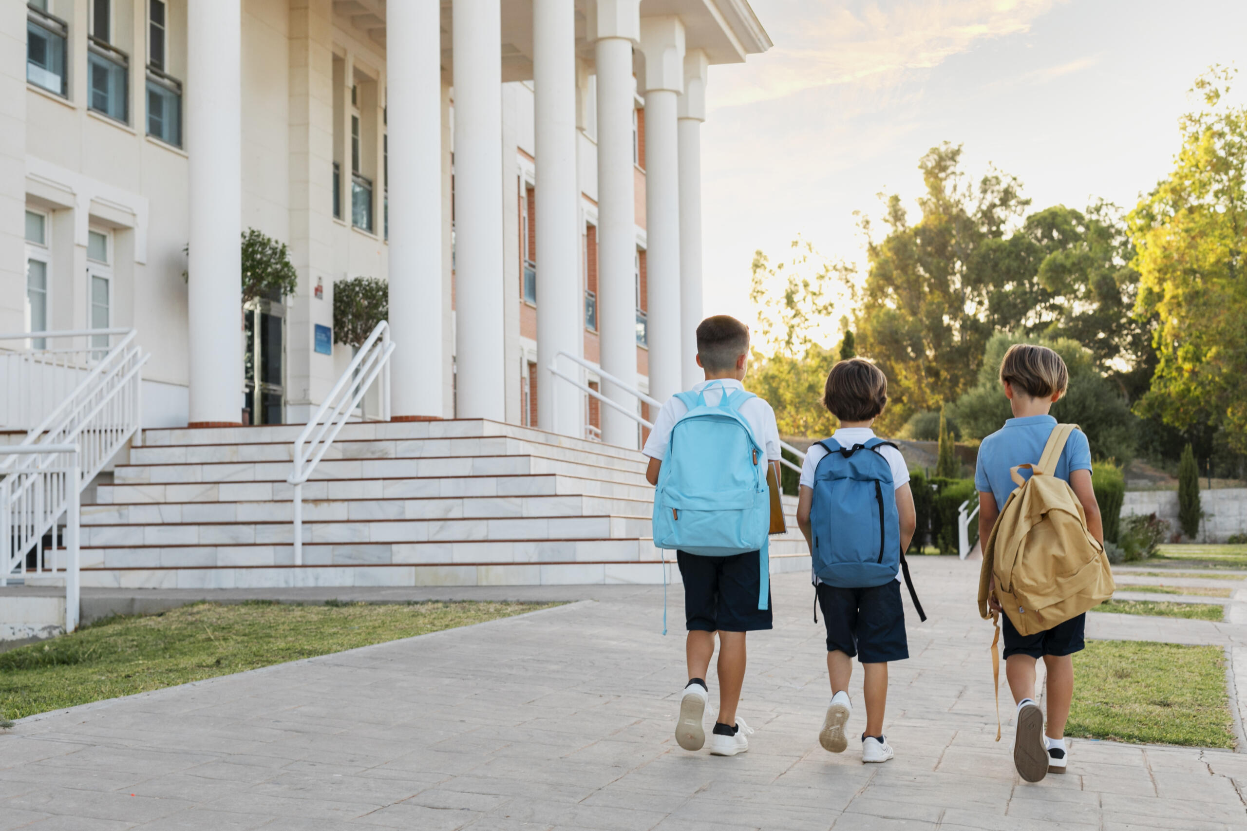 tres niños con sus mochilas de espaldas a la cámara, caminando hacia la entrada del colegio