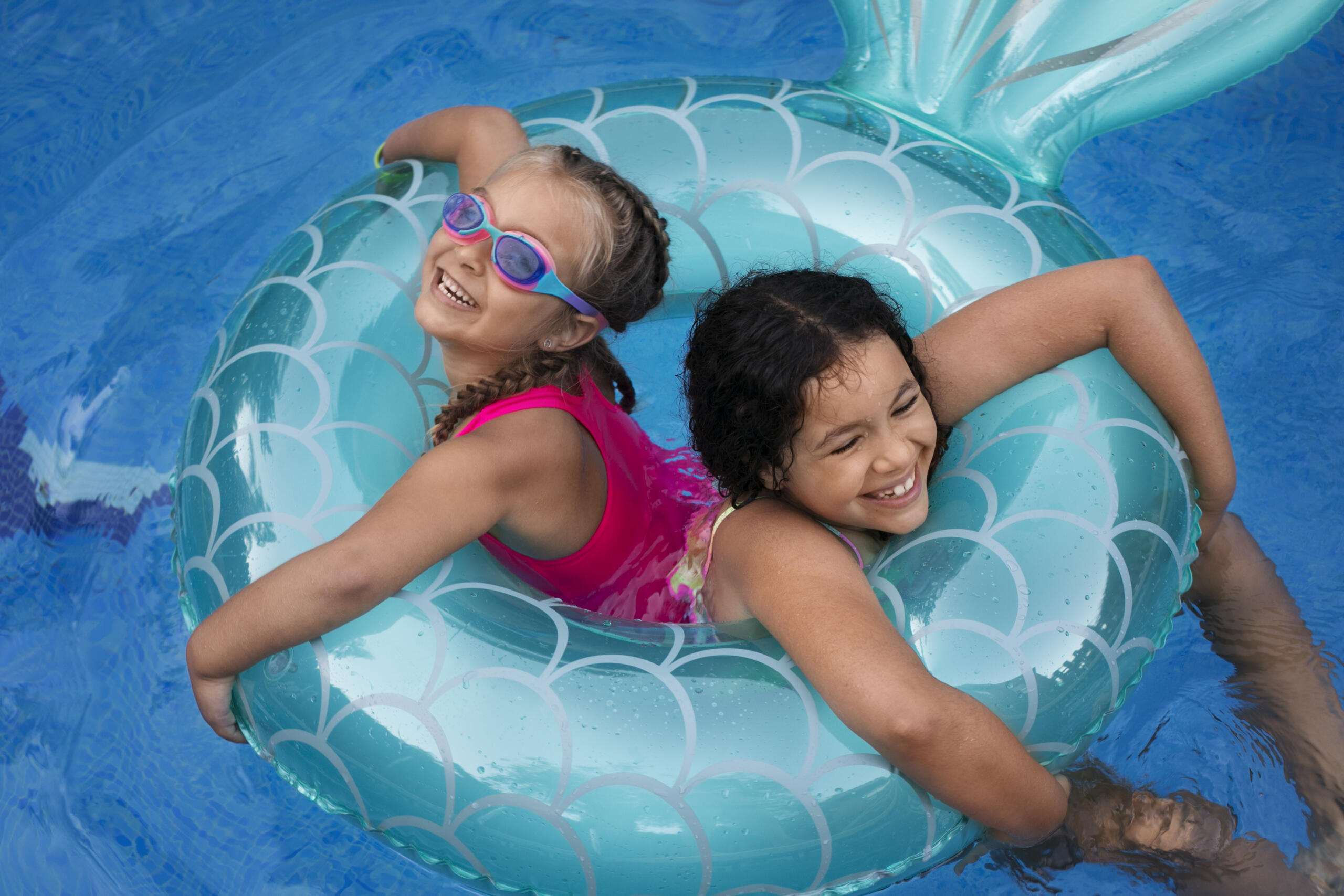 toma contrapicada de dos niñas sonrientes en medio de un flotador redondo con cola de sirena dentro de la piscina