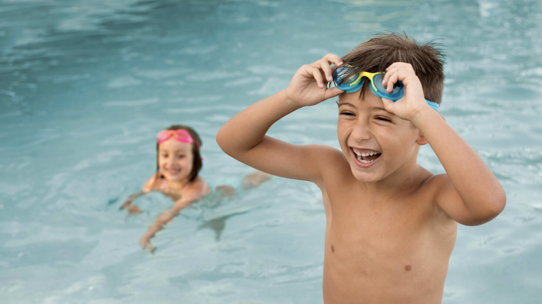 niño en primer plano sonriente colocandose las gafas de natacion con vista de niña en el fondo ya metida en la piscina