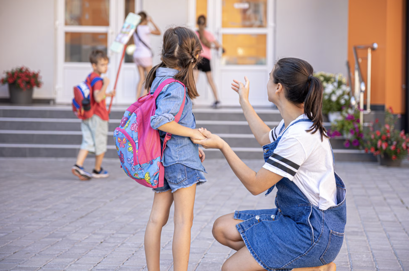 mamá en cunclillas junto a su hija señalándole el ingreso de la escuela mientras se ve a otros niños en fondo desenfocado