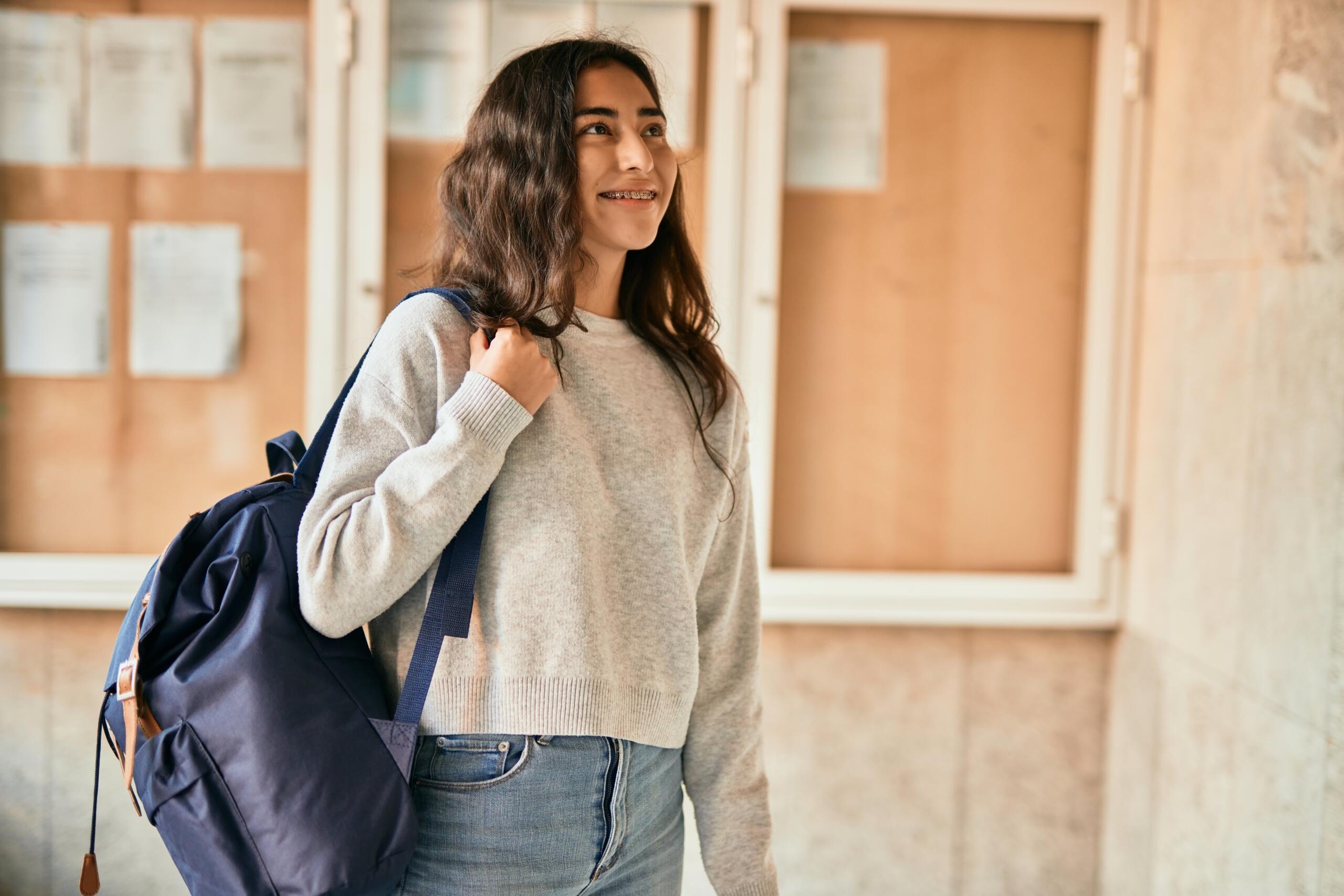 chica de secundaria mirando sonriente un pasillo de universidad sosteniendo su mochila en un hombro