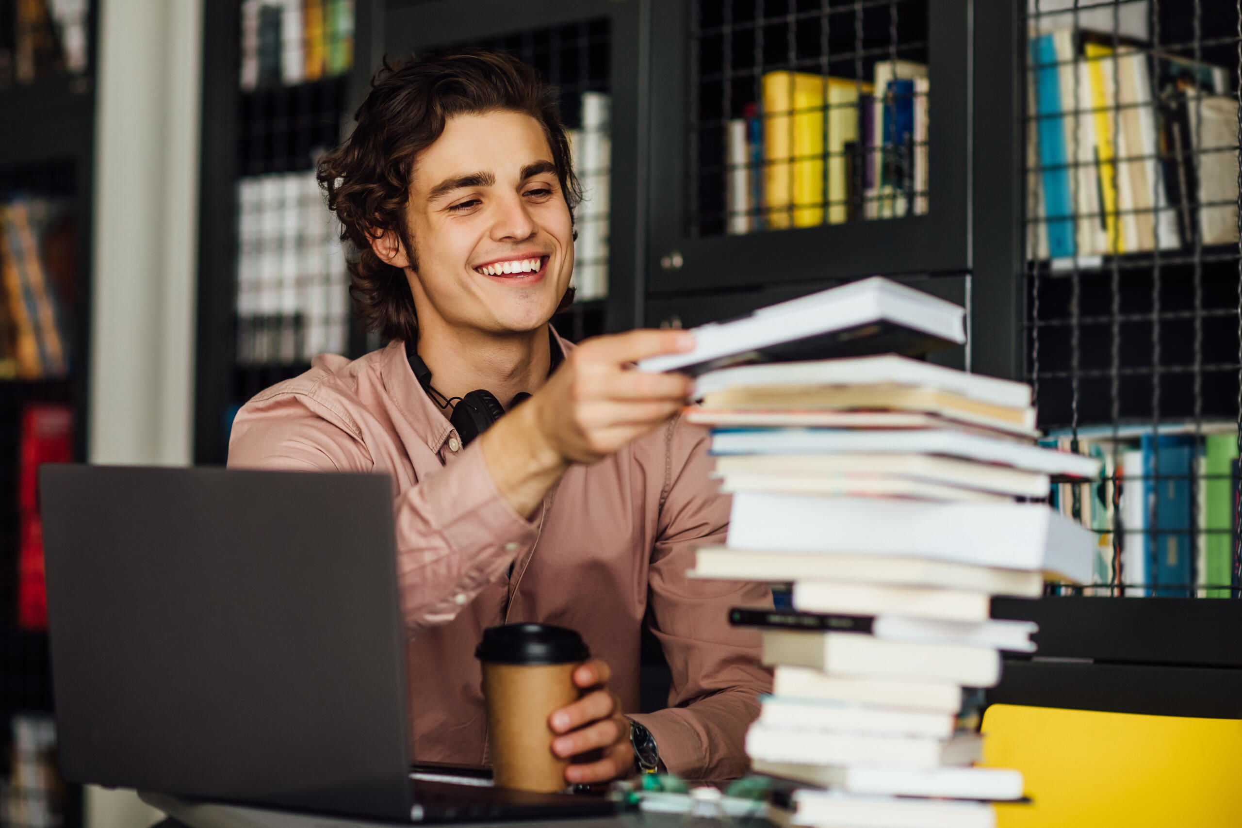 chico sonriente apilando una torre de libros en su escritorio mientras estudia en una biblioteca